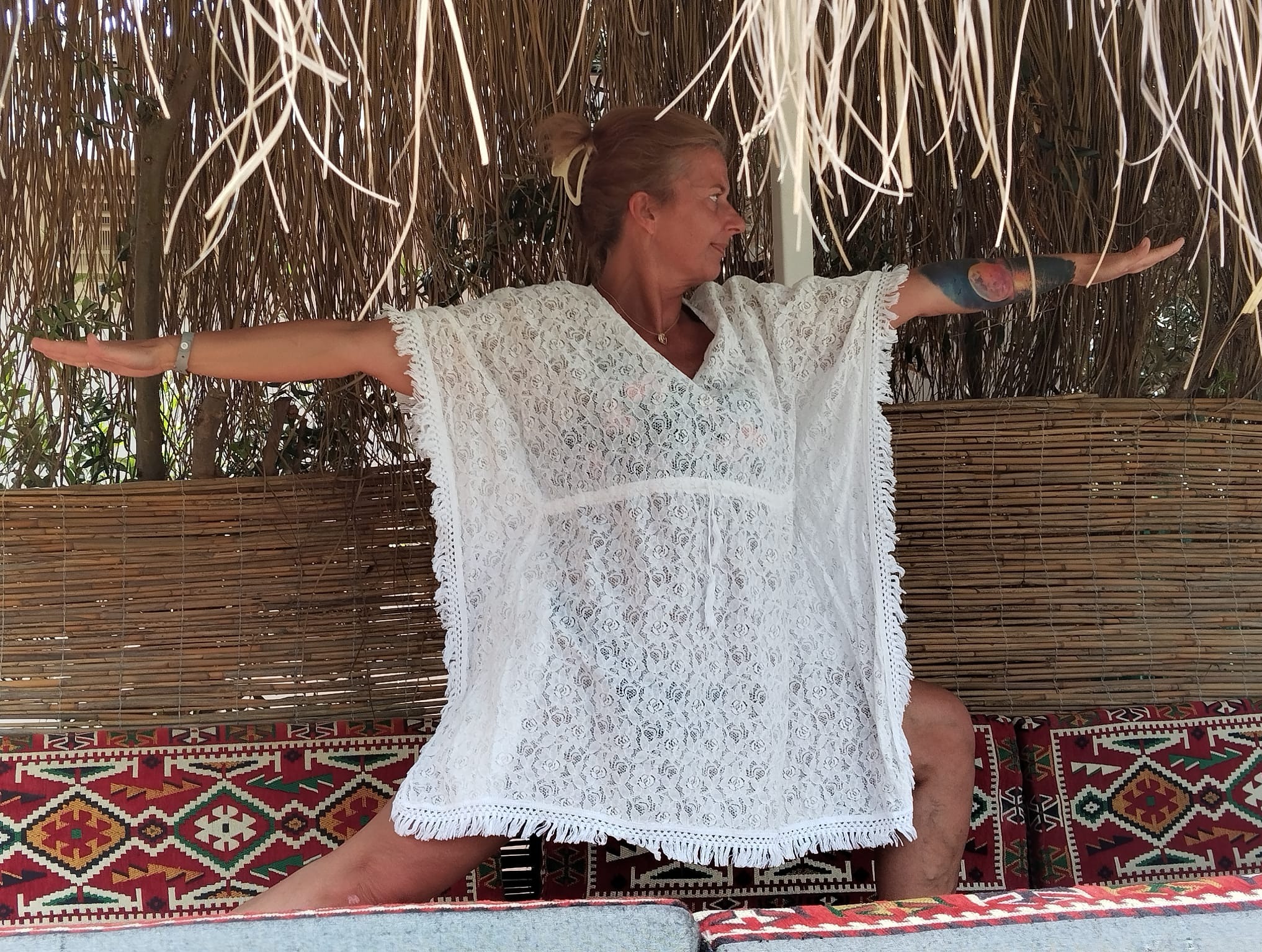 Kobieta w białej tunice wykonuje asanę jogi w plenerze, na tle bambusowej ściany i wzorzystych poduszek. Widoczny tatuaż na ramieniu.