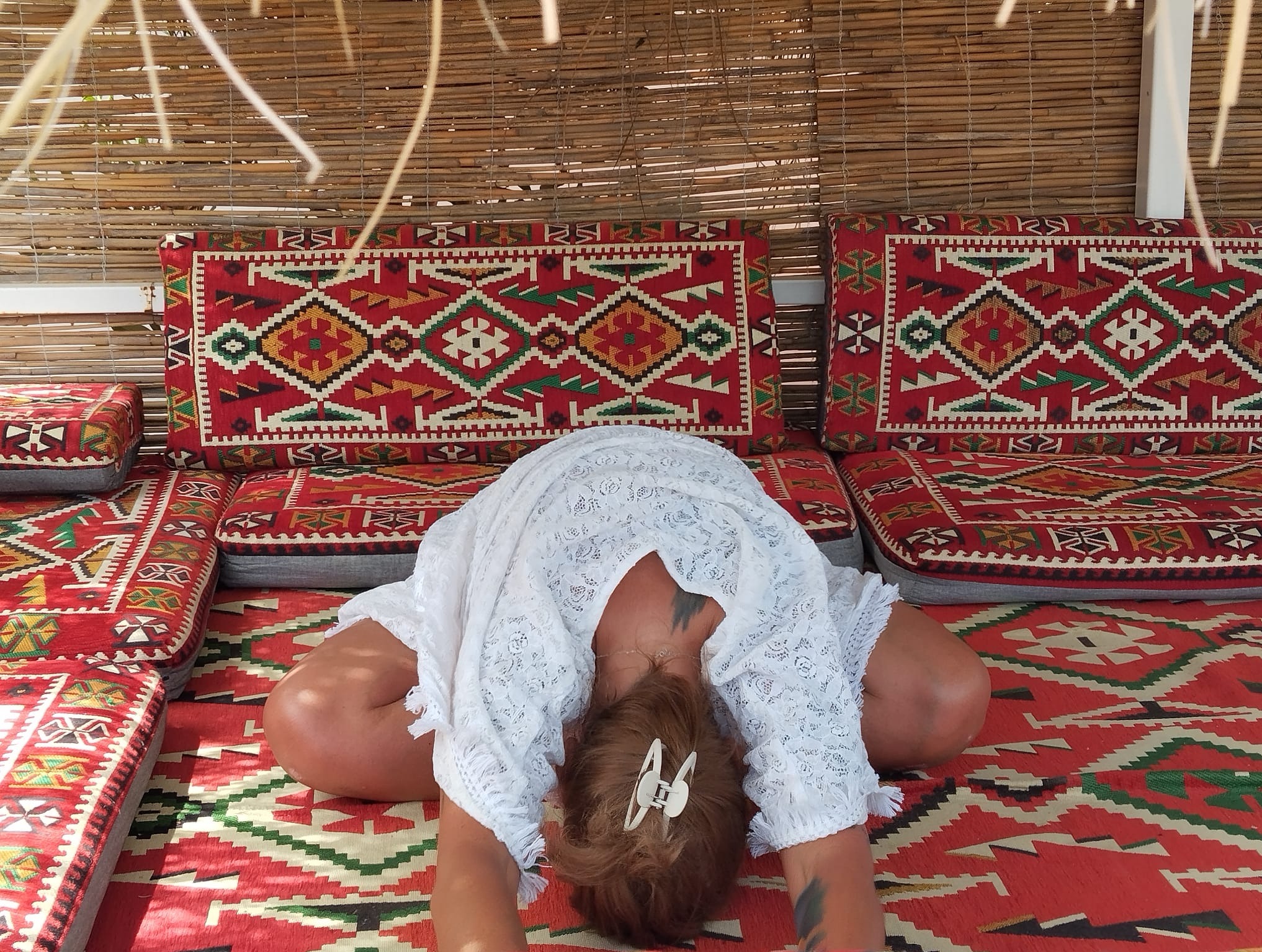 Kobieta w białej sukience wykonuje skłon do przodu w pozycji siedzącej na wzorzystym dywanie i poduszkach w orientalnym stylu, pod bambusowym zadaszeniem.