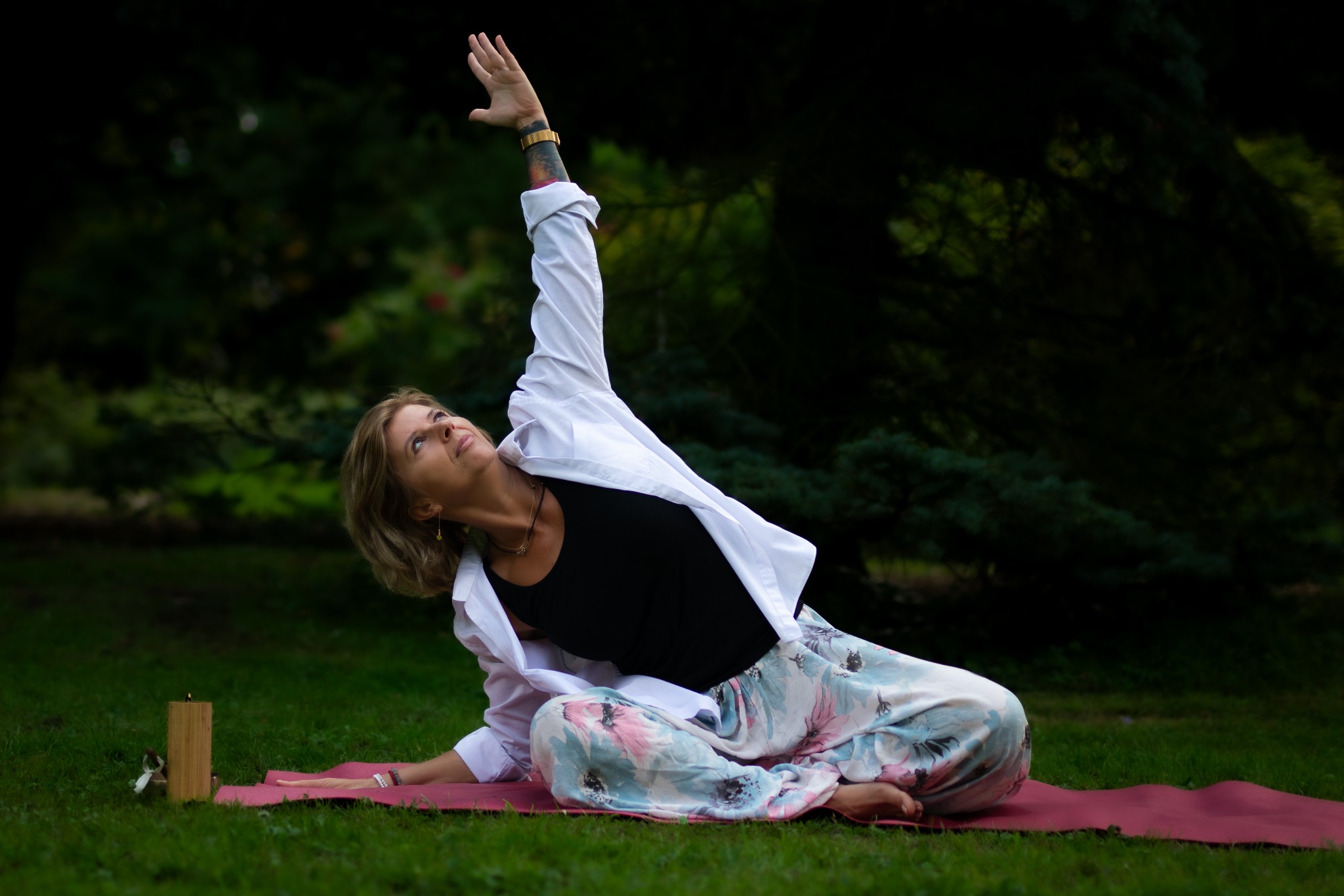 Kobieta w kwiecistych spodniach i białej koszuli wykonuje pozycję jogi na czerwonej macie w plenerze. Widoczny tatuaż na ramieniu i drewniany element dekoracyjny.