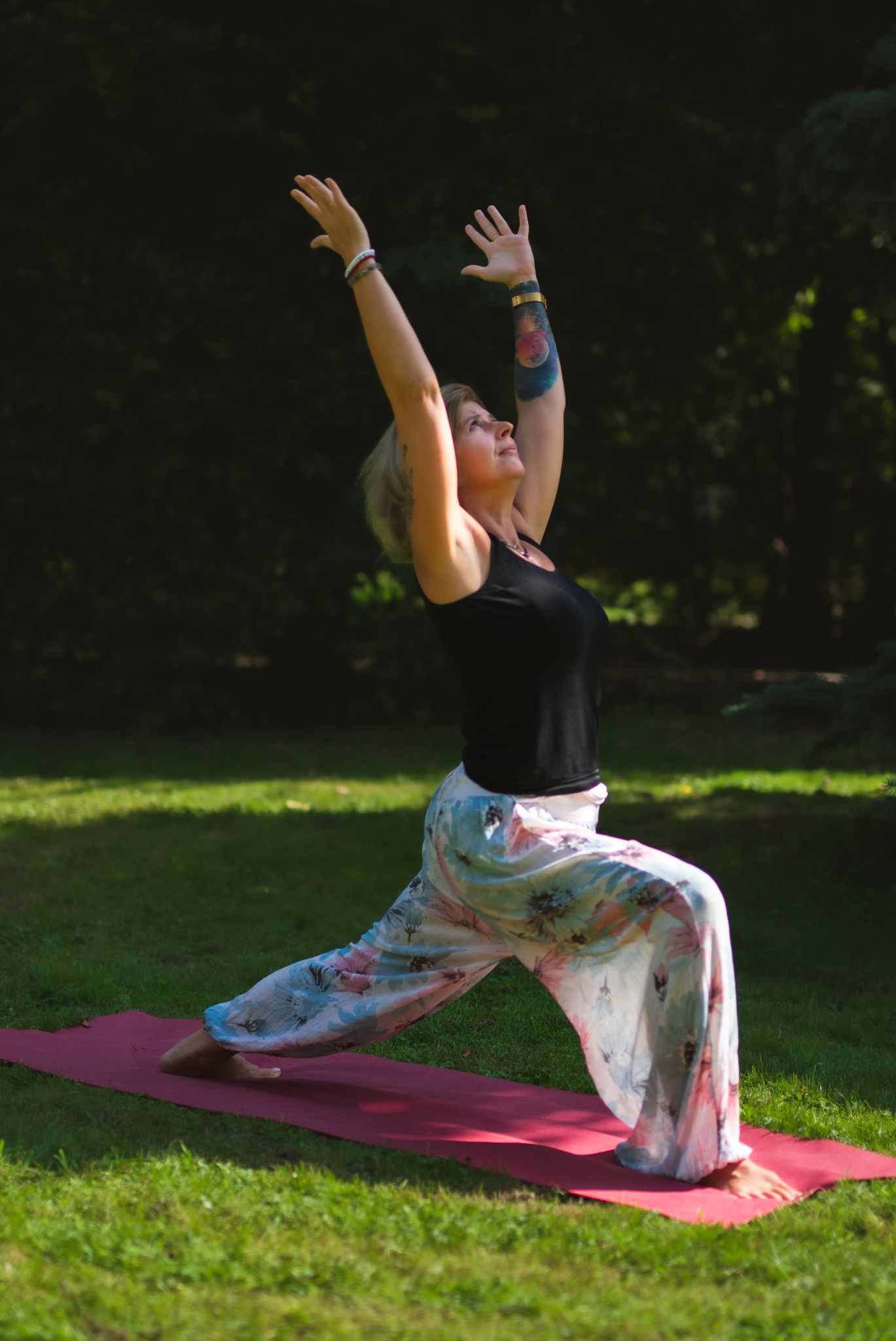 Kobieta w pozycji wojownika I na macie do jogi w plenerze, z tatuażem na ramieniu i podniesionymi rękami. Ubrana w czarny top i wzorzyste spodnie.