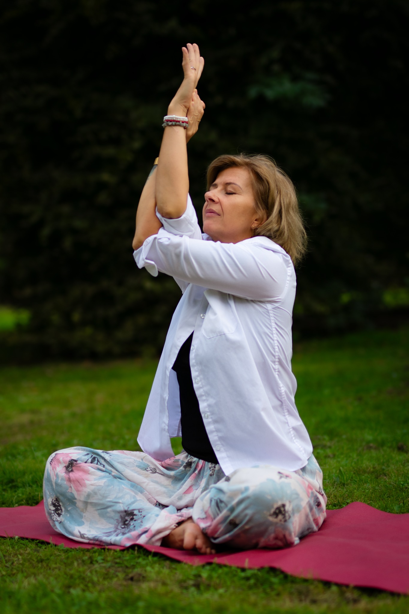 Kobieta w białej koszuli i kwiecistych spodniach siedzi na czerwonej macie na trawie, wykonując pozycję jogi z rękami uniesionymi nad głową na tle zieleni.
