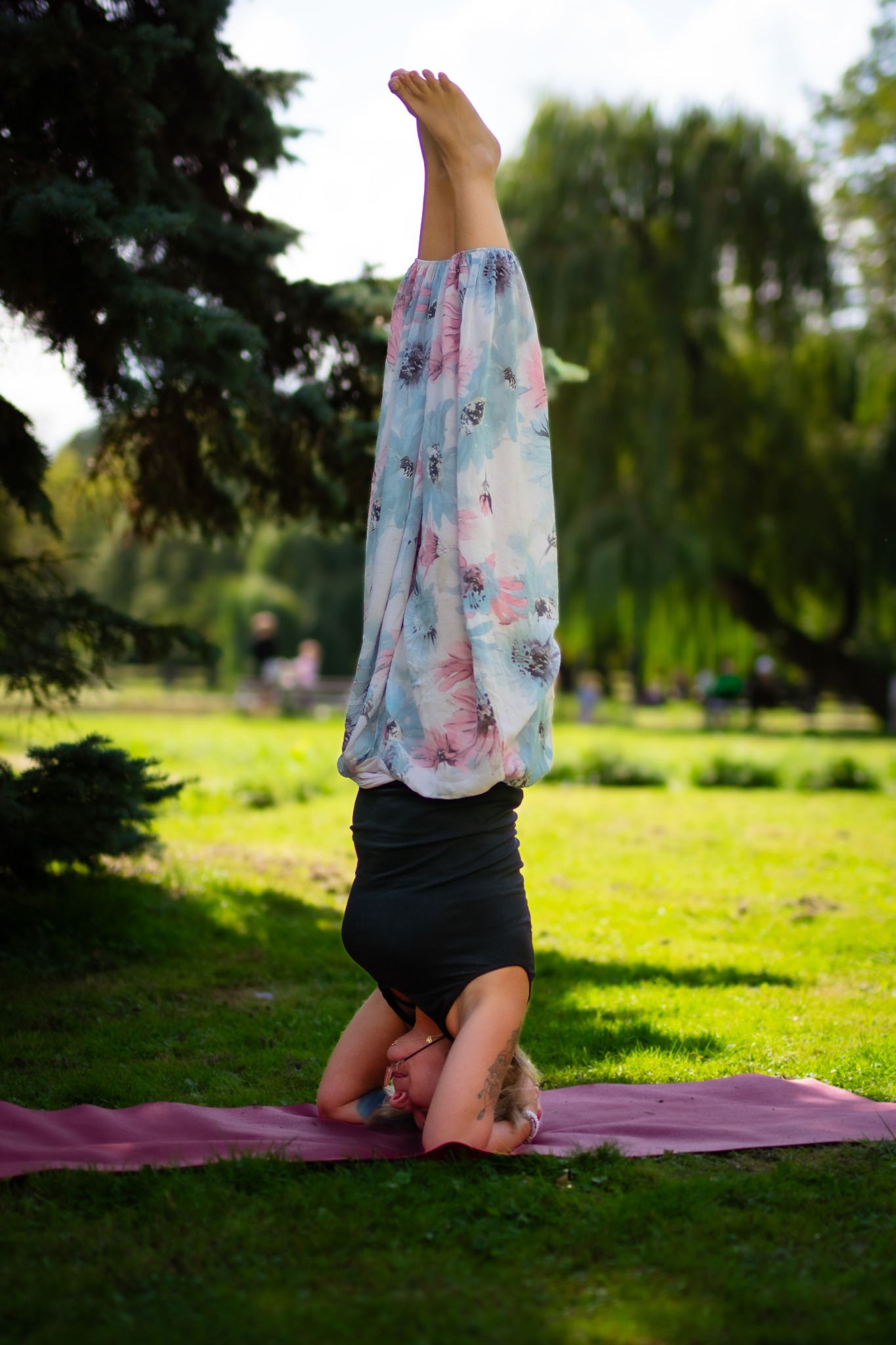 Kobieta w pozycji stania na głowie na macie do jogi w parku. Ubrana w czarny top i kwieciste spodnie. Widoczne drzewa i trawa w tle, miękkie światło.