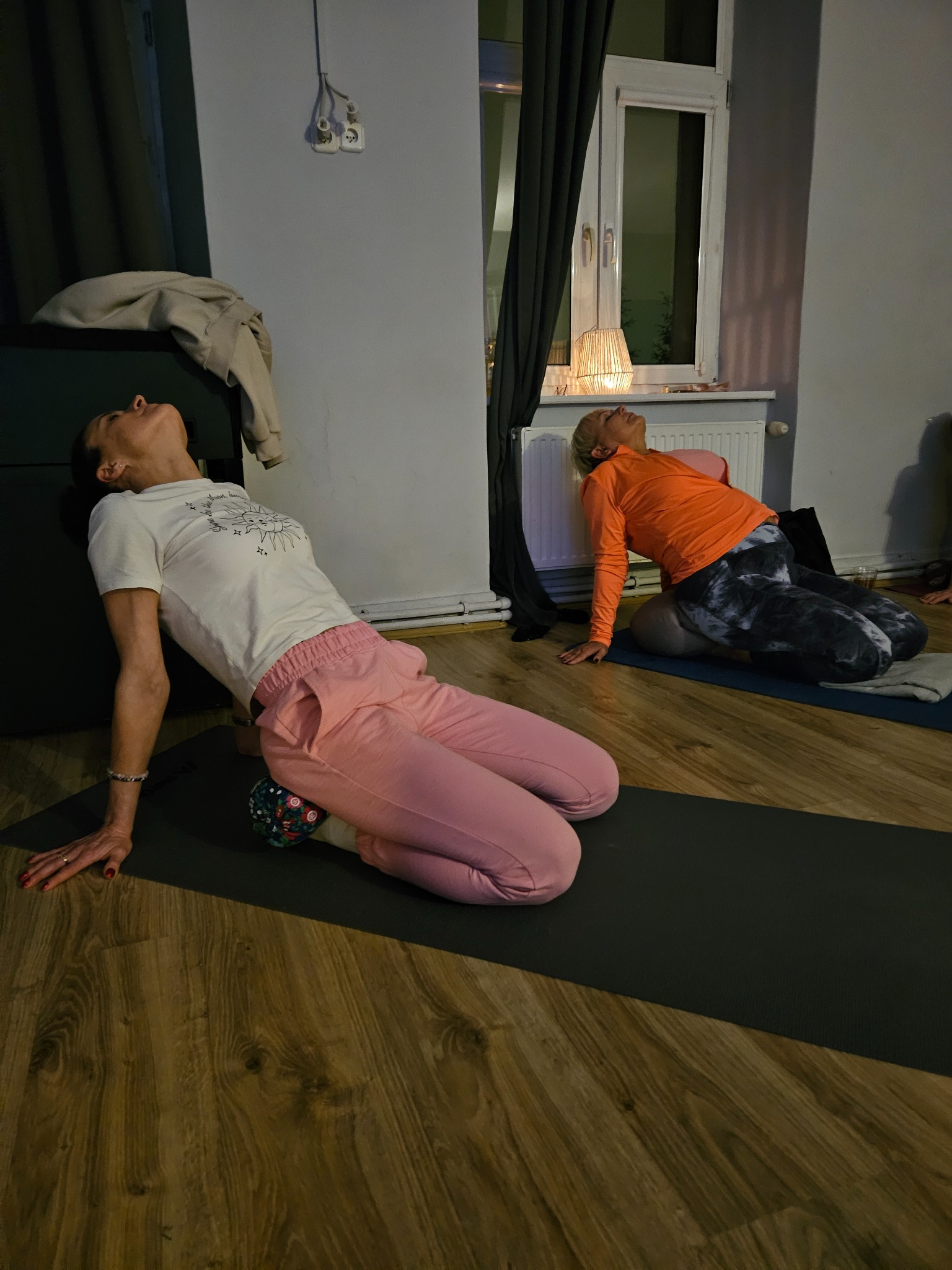 Dwie kobiety w pozycji odchylenia do tyłu na matach do ćwiczeń w jasnym pomieszczeniu. Jedna z wałkiem pod kolanami. Widoczne okno i grzejnik.