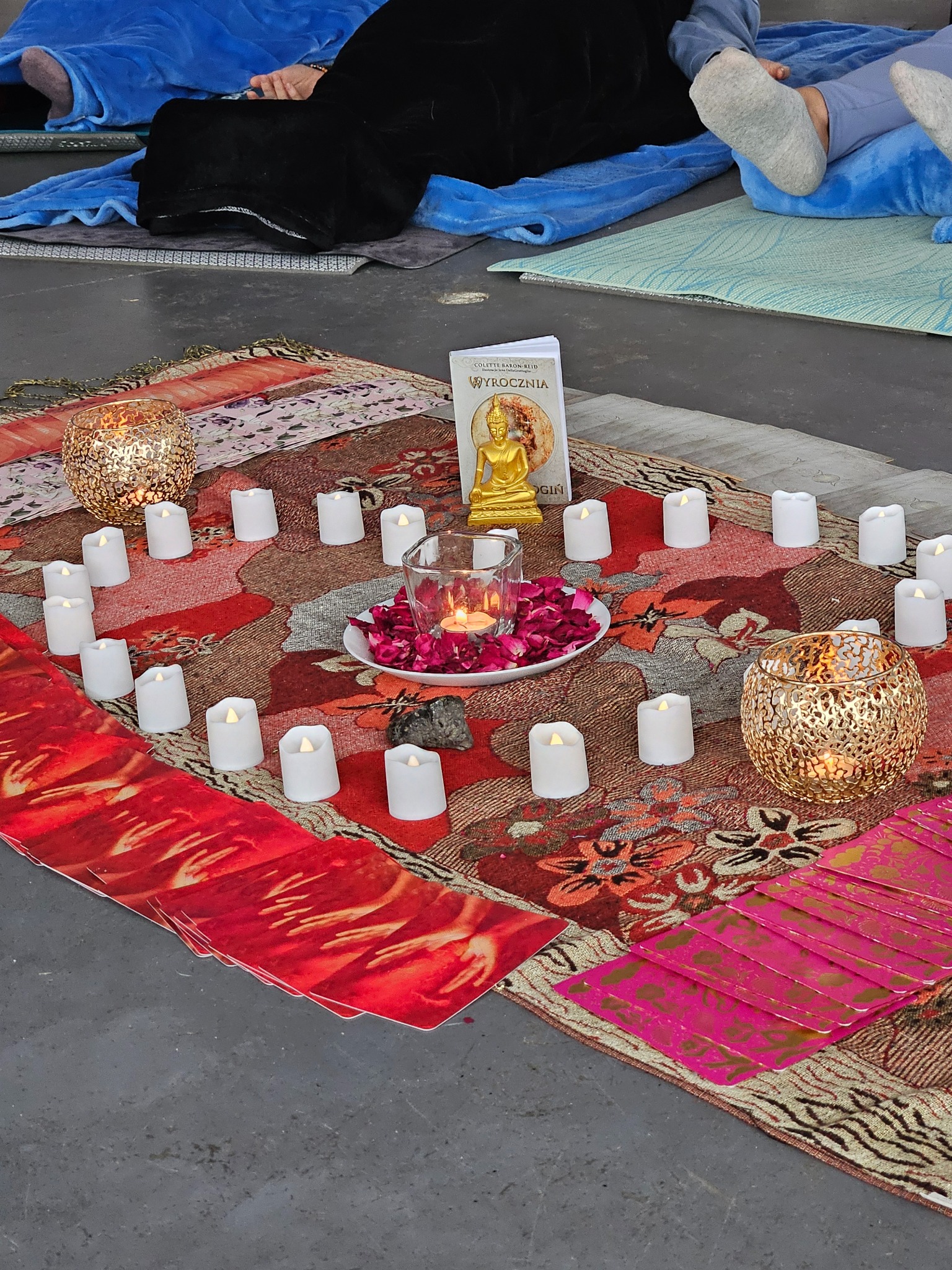 Dekoracja z świec, płatków róż i figurki Buddy na wzorzystym dywanie, tworząca atmosferę relaksu podczas zajęć jogi lub medytacji. Widoczne maty do ćwiczeń.