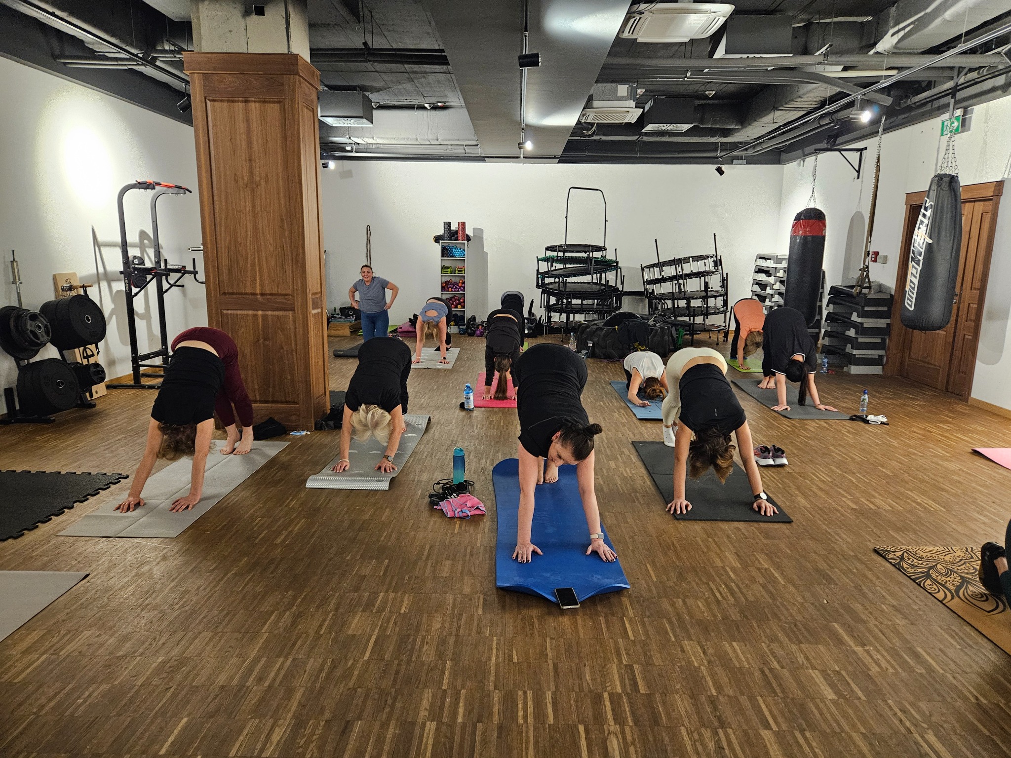 Grupa osób ćwiczy jogę w przestronnym, jasnym pomieszczeniu z drewnianą podłogą. Widoczne maty do ćwiczeń, sprzęt sportowy i worki bokserskie w tle.