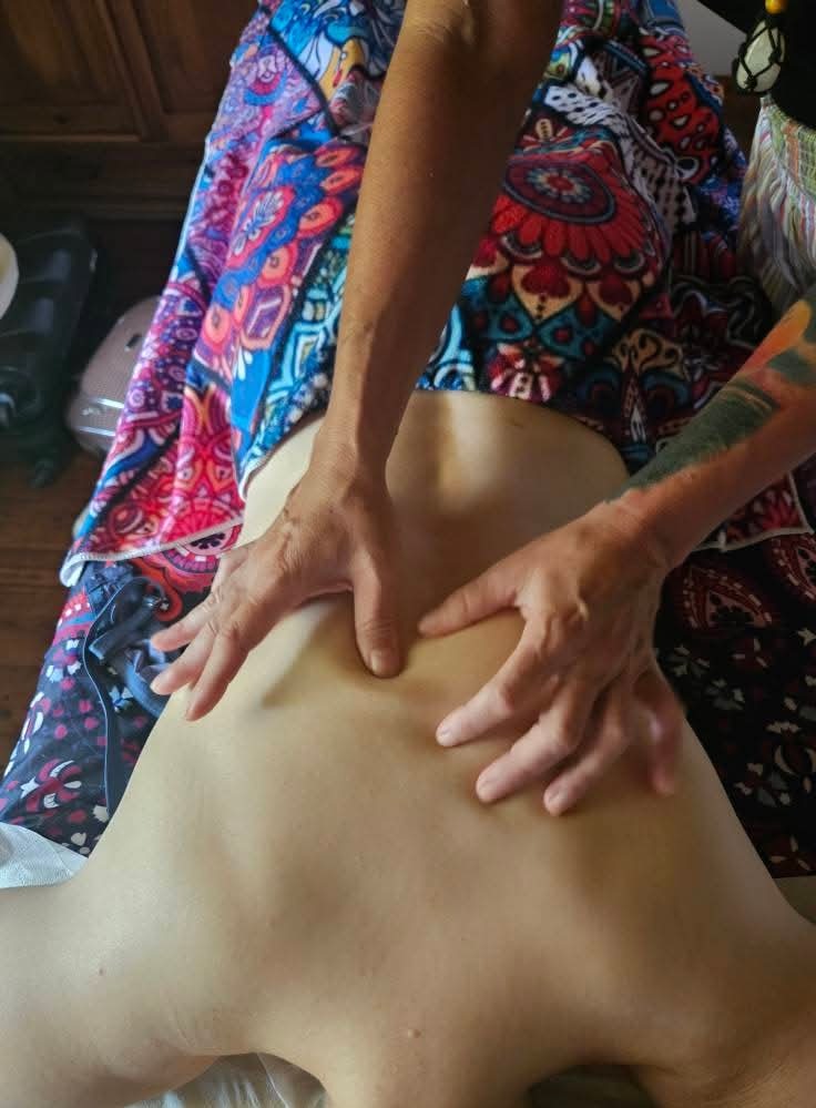 Masażysta uciska dłońmi plecy klienta leżącego na stole do masażu, okrytego kolorową tkaniną. Widoczne tatuaże na ramieniu masażysty.