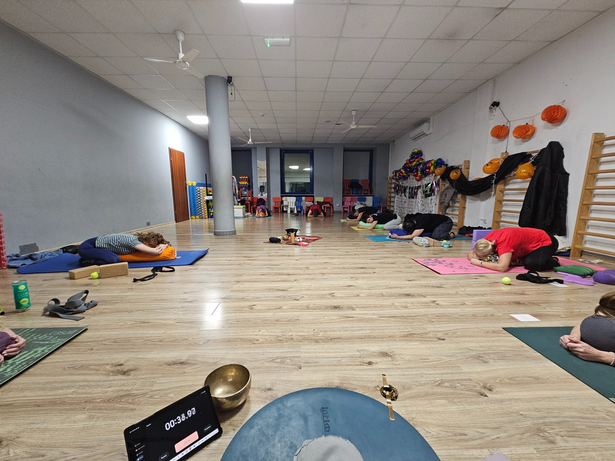 Grupa osób w pozycji relaksacyjnej na matach do jogi w przestronnym, jasnym pomieszczeniu z dekoracjami halloweenowymi w tle.