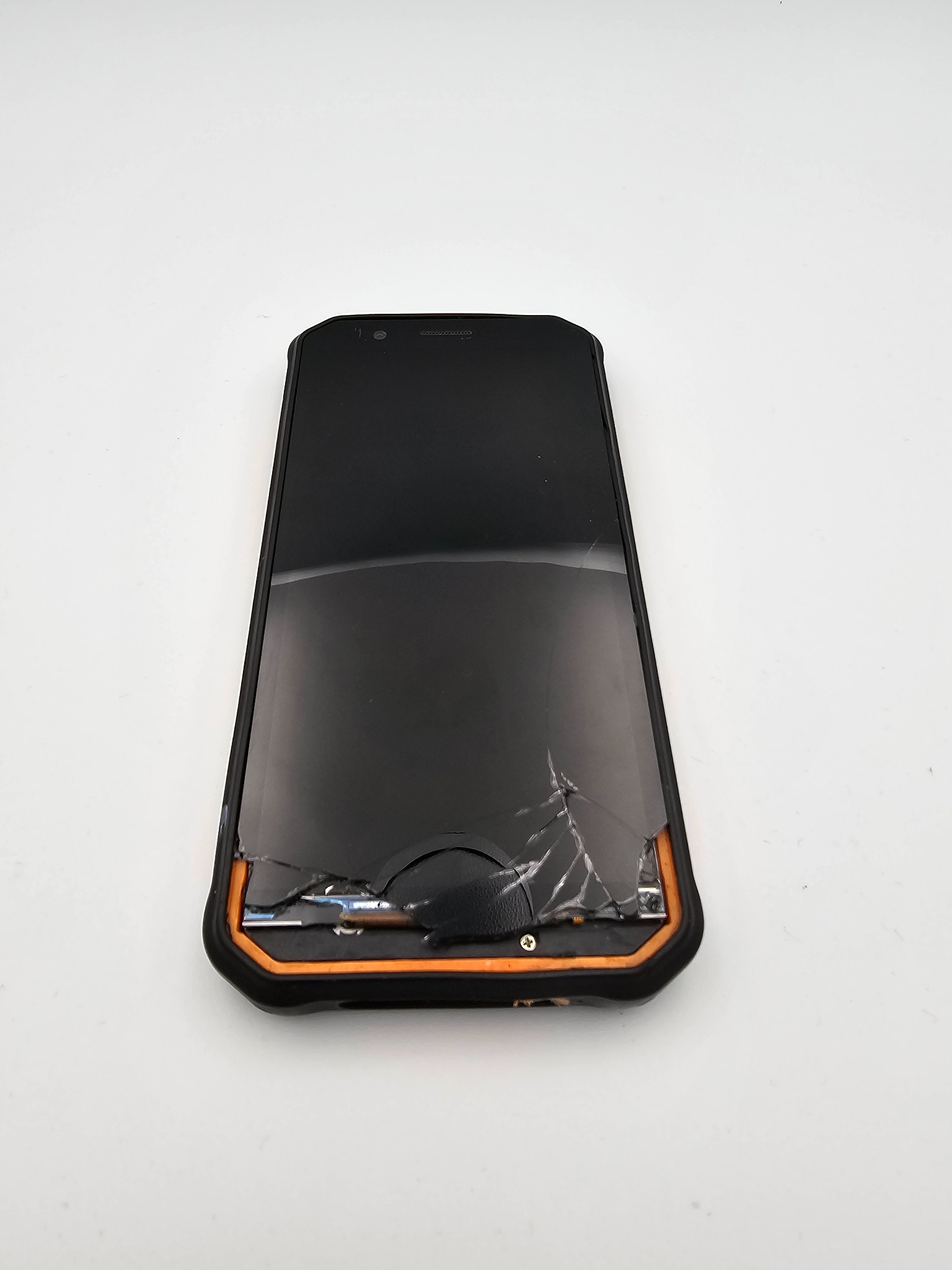 Uszkodzony smartfon z pękniętym ekranem, widoczne liczne rysy i uszkodzenia. Obudowa w kolorze czarnym z pomarańczowymi akcentami. Białe tło.