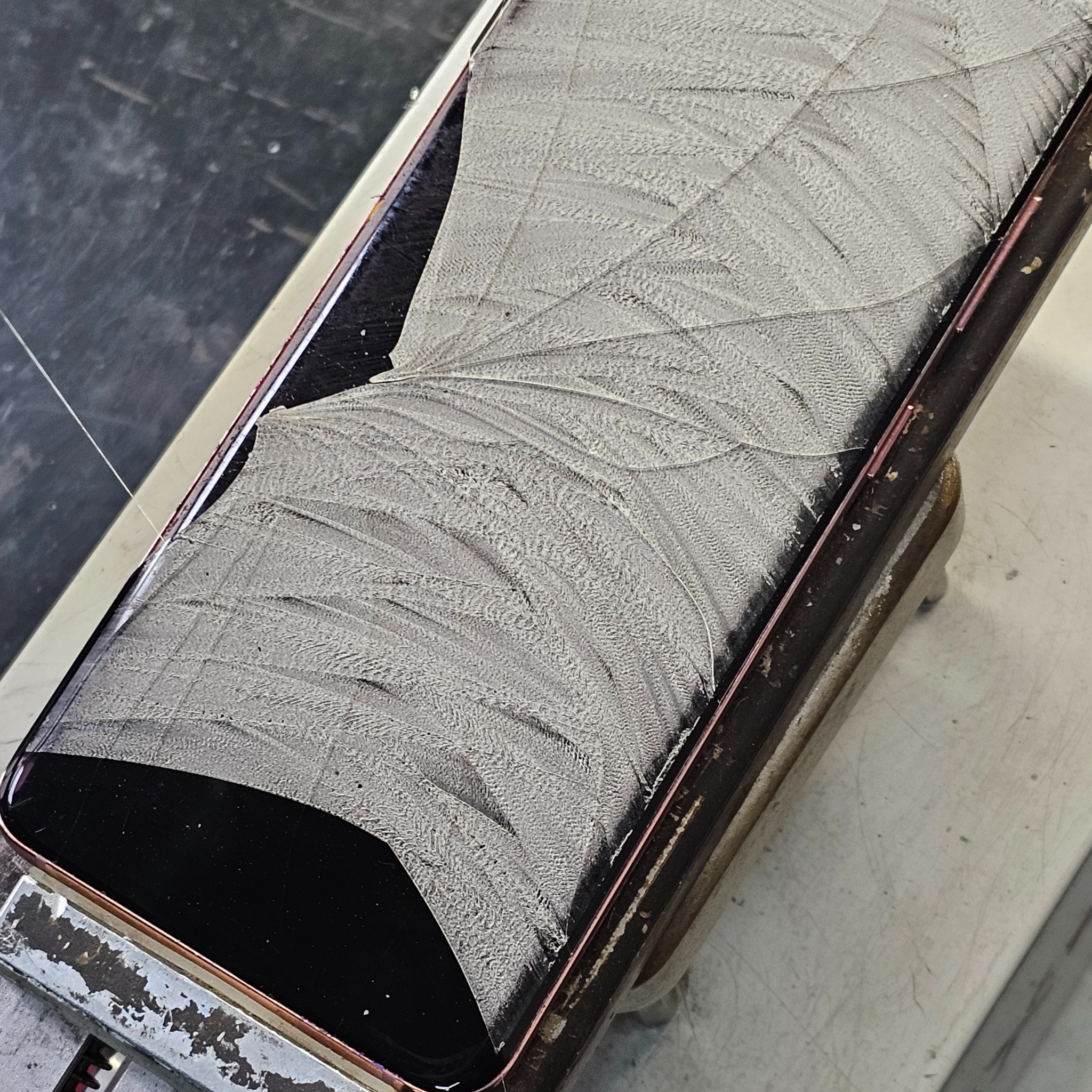 Uszkodzony ekran smartfona z widocznymi pęknięciami na całej powierzchni, leżący na stole warsztatowym. Widoczne narzędzia serwisowe w tle.