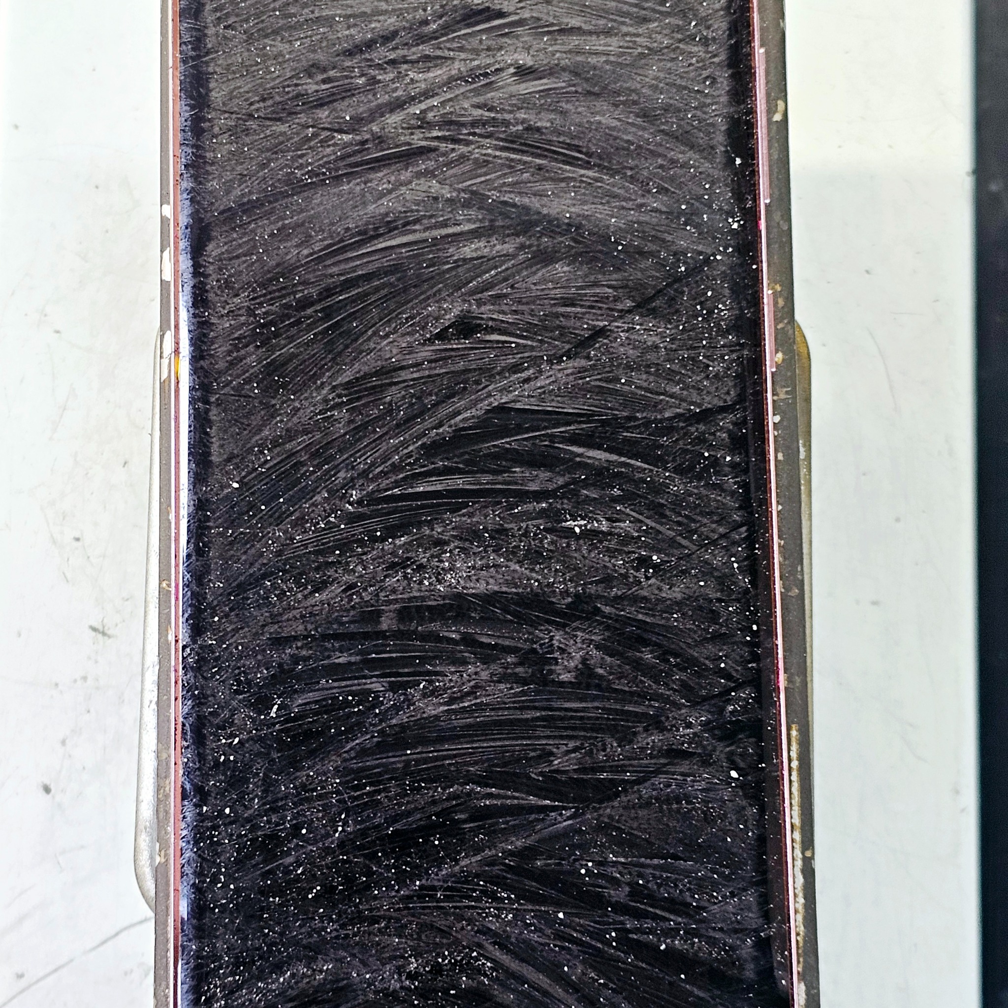 Ekran smartfona po czyszczeniu, widoczne smugi i drobinki kurzu. Krawędź obudowy zarysowana, tło białe. Detal po naprawie w serwisie.