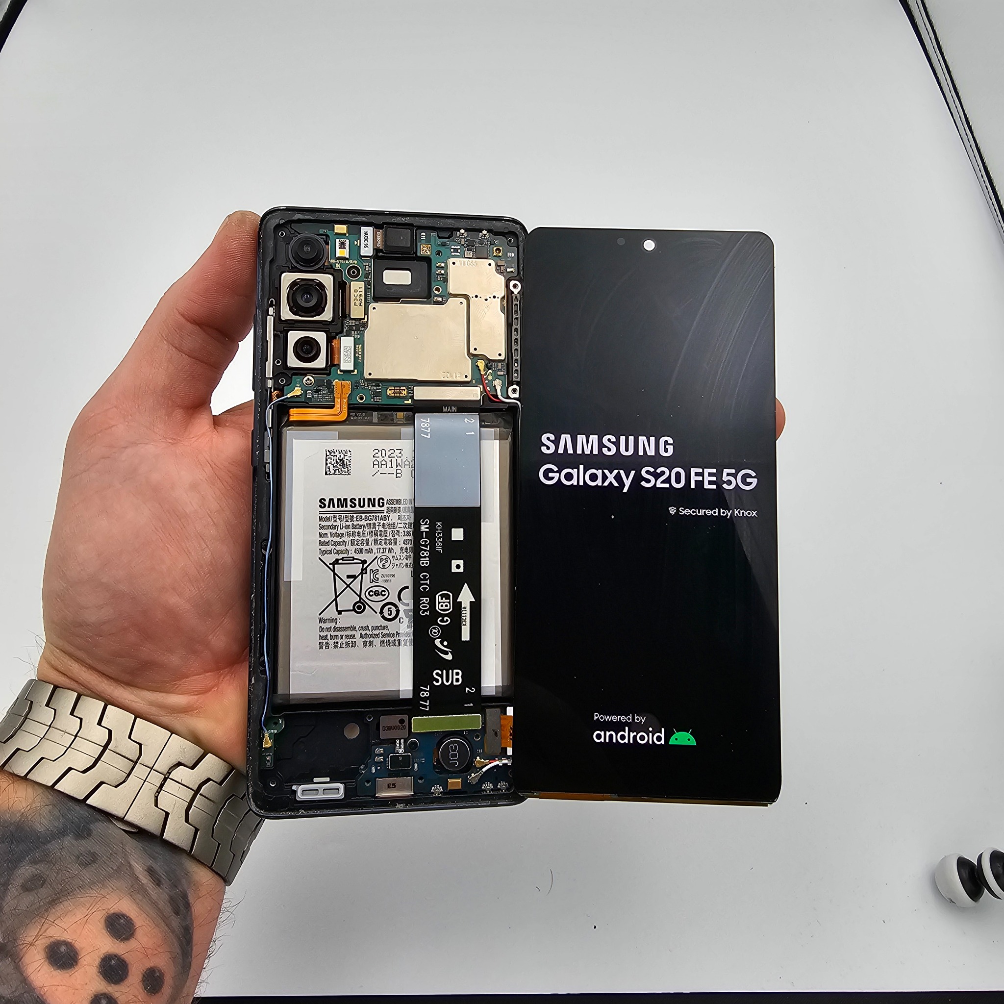 Otwarty smartfon Samsung Galaxy S20 FE 5G w dłoni, ukazujący wewnętrzne komponenty i baterię. Widoczny ekran z logo Android. Precyzyjna naprawa elektroniki.