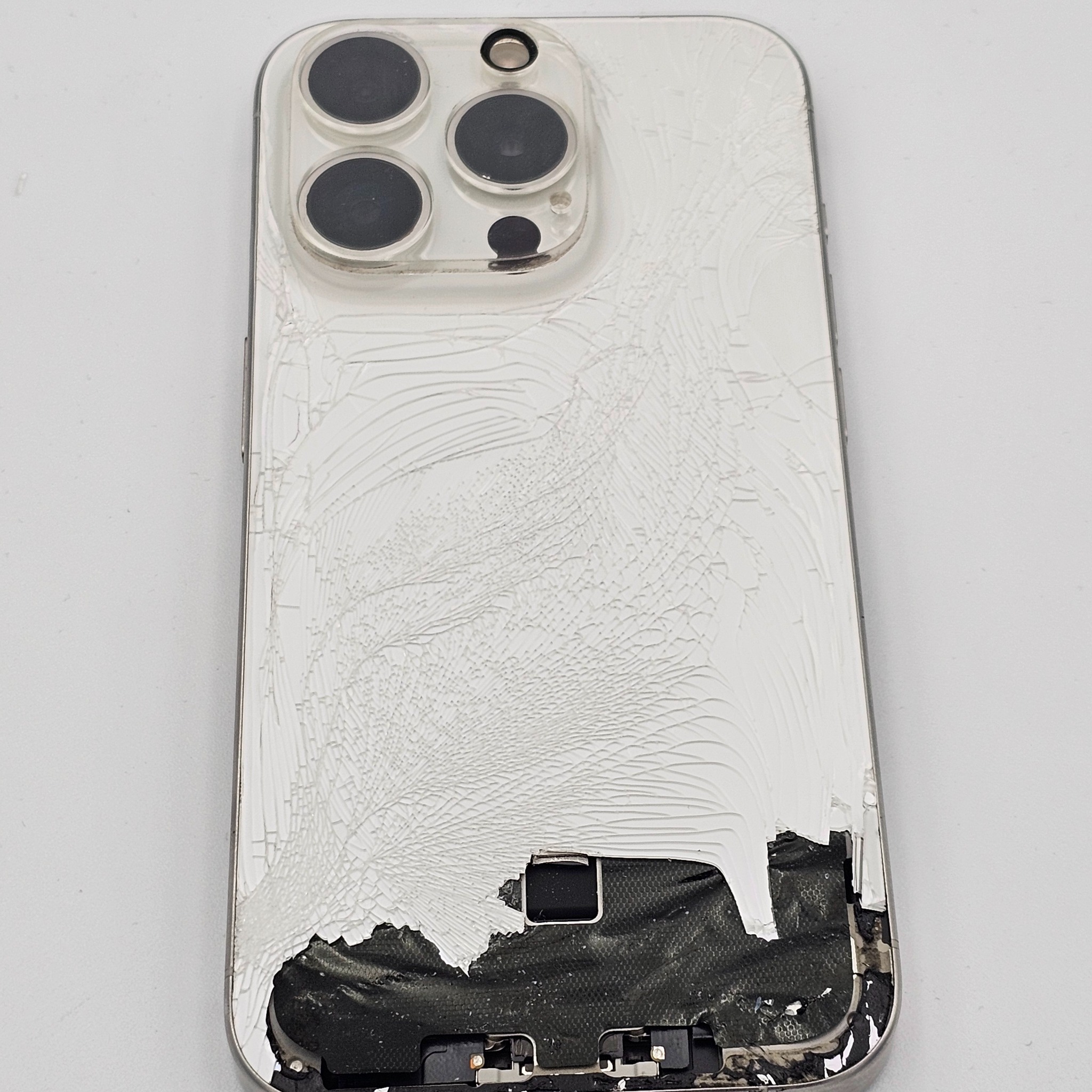 Uszkodzony, biały iPhone z pękniętą obudową i widocznymi elementami wewnętrznymi. Widoczne trzy obiektywy aparatu. Tło białe, jednolite.