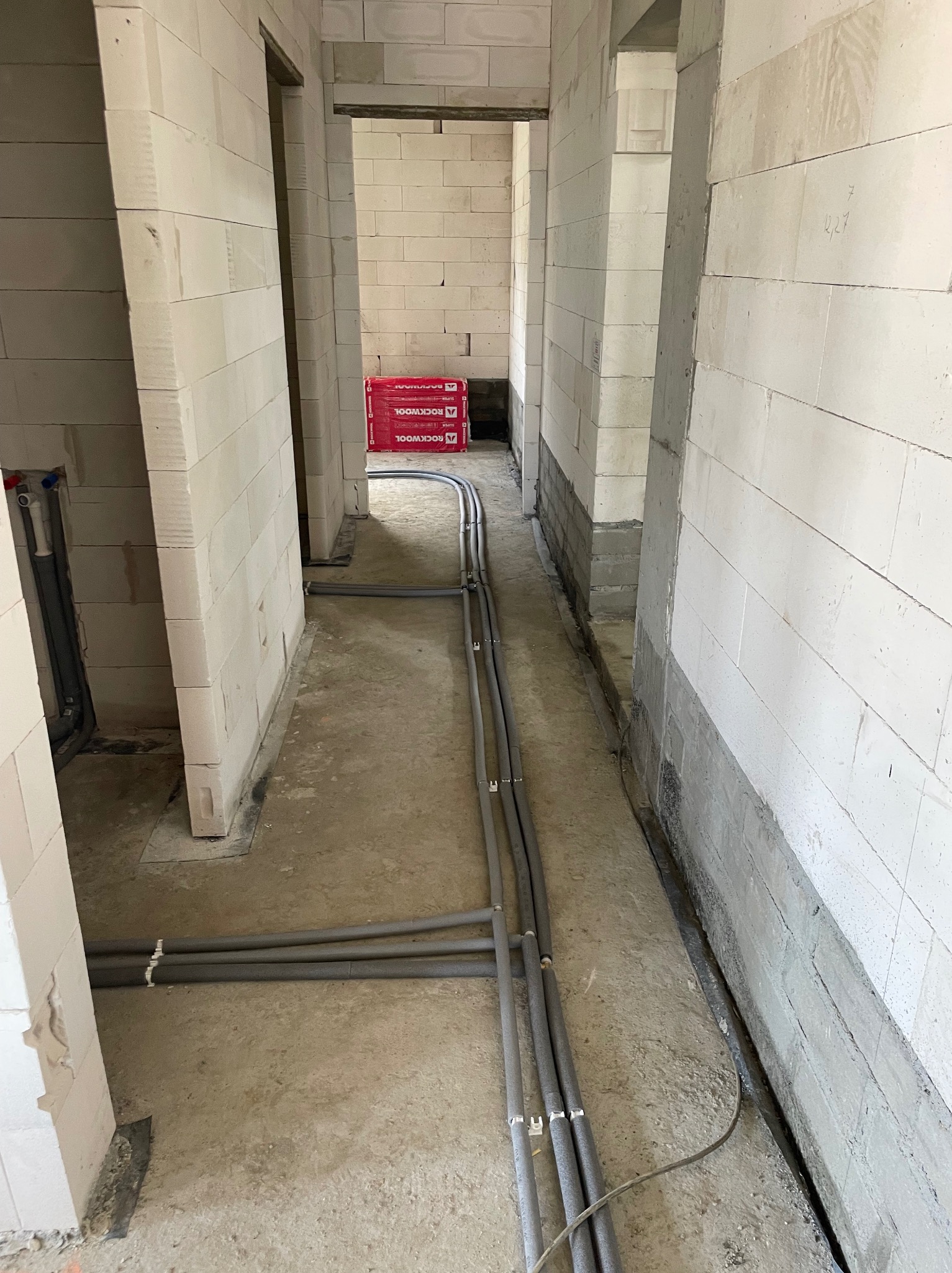 Szare rury instalacji hydraulicznej ułożone na surowym podłożu w budynku z białych bloczków. Widoczne fragmenty ścian i otwory drzwiowe. W tle paczki wełny mineralnej.
