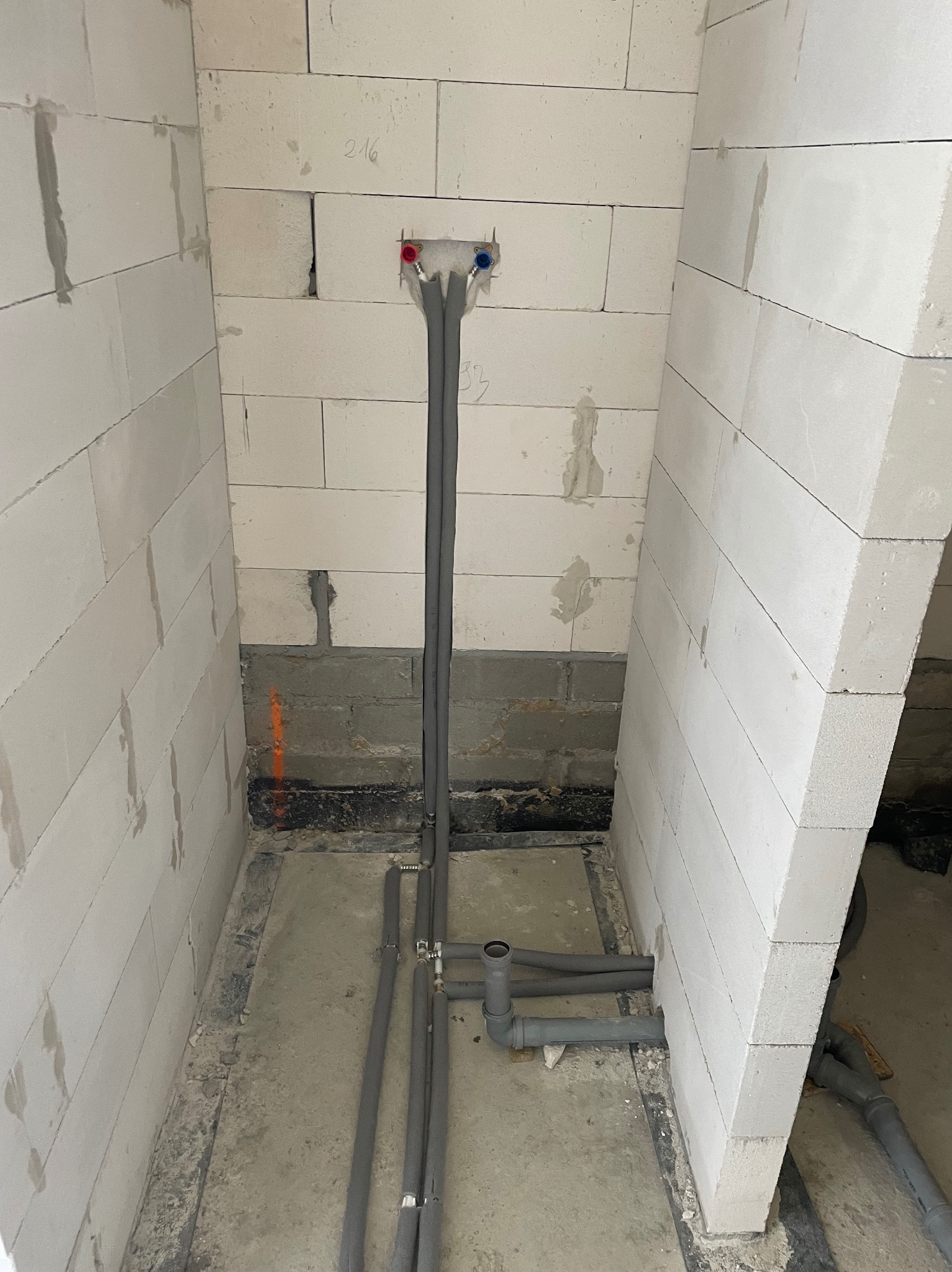 Szare rury instalacji wodnej z czerwonym i niebieskim zaworem, prowadzone w surowym pomieszczeniu z białych bloczków. Widoczne fragmenty podłogi i ściany w trakcie prac hydraulicznych.