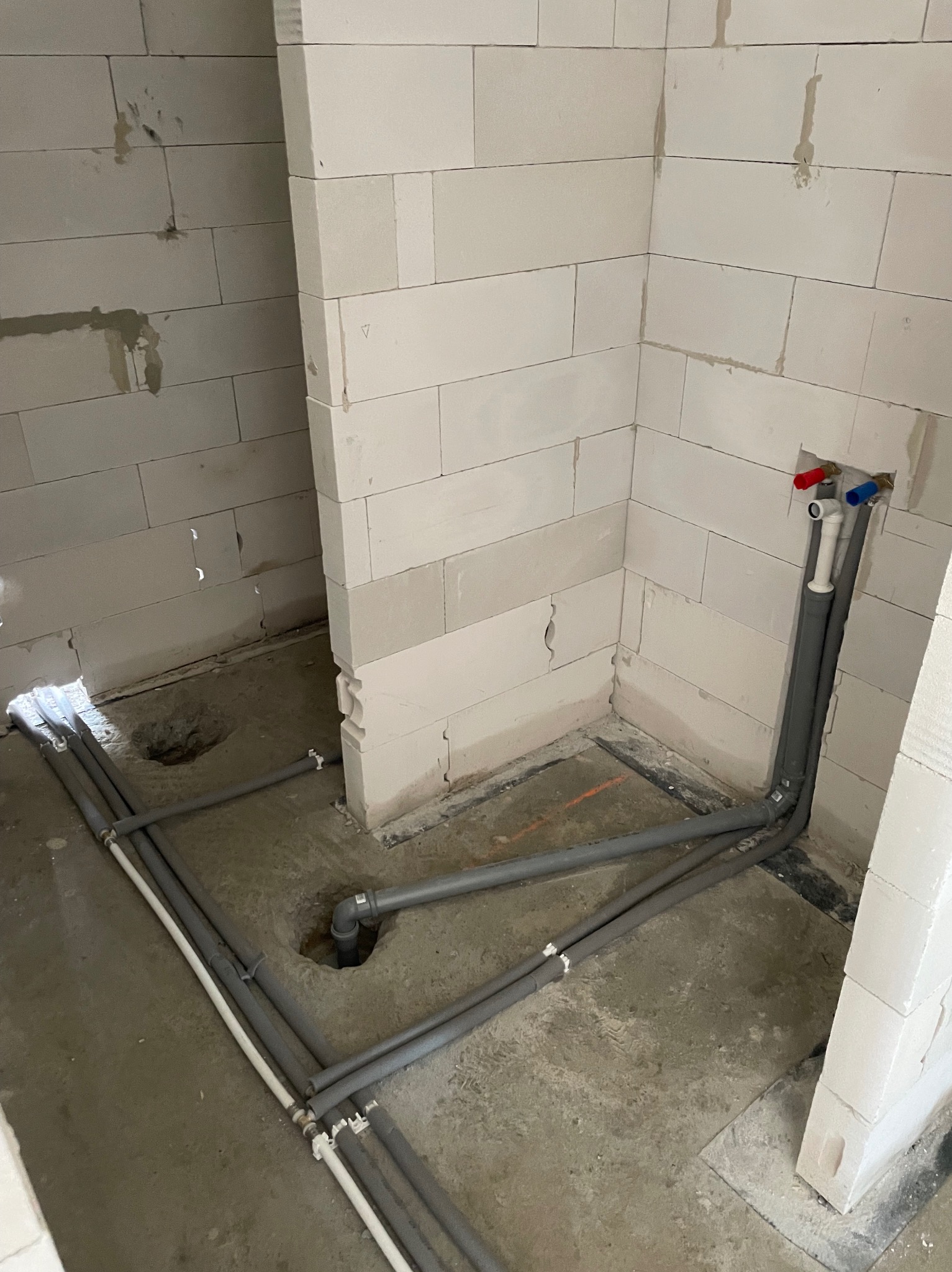 Szare i białe rury instalacji wodnej ułożone na surowym betonie w rogu pomieszczenia z białych bloczków, z widocznymi otworami w podłodze i wystającymi zaworami.