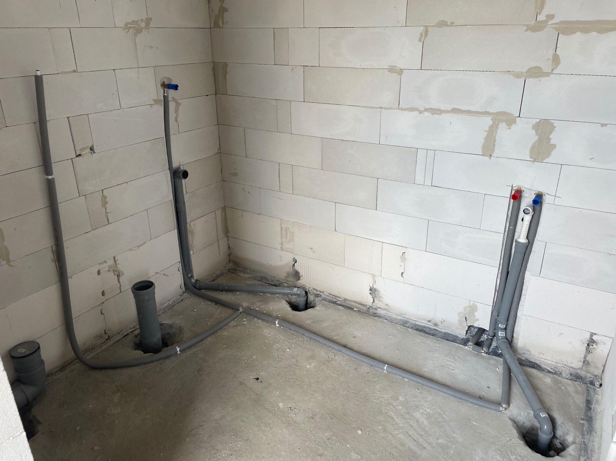 Szare rury kanalizacyjne i wodne w surowej łazience z białymi ścianami z betonu komórkowego. Widoczne otwory w podłodze i wystające zawory.