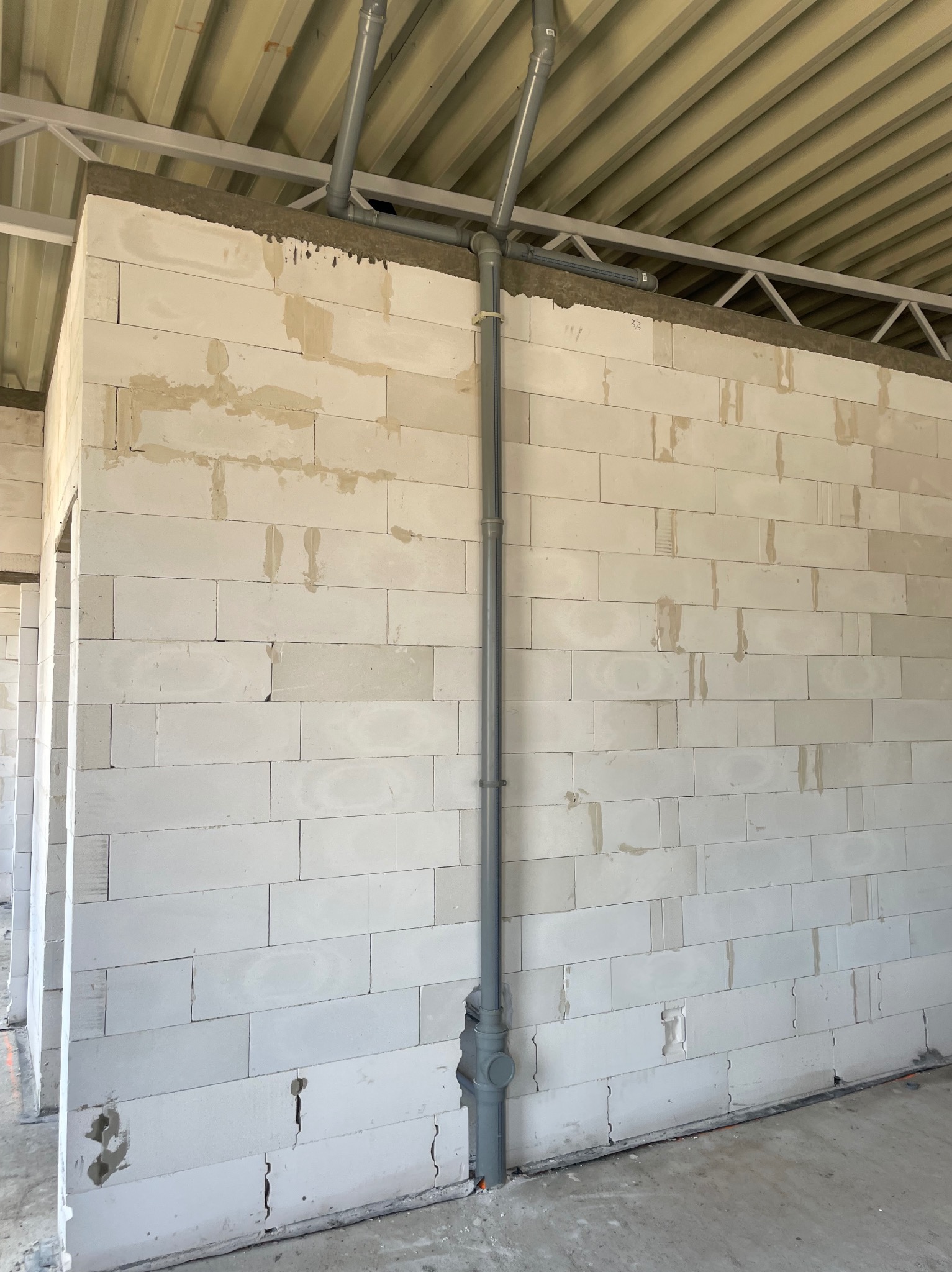 Szara rura pionowa z rozgałęzieniami przy ścianie z białych bloczków. Instalacja wodno-kanalizacyjna w surowym stanie, widoczne elementy konstrukcji stropu.