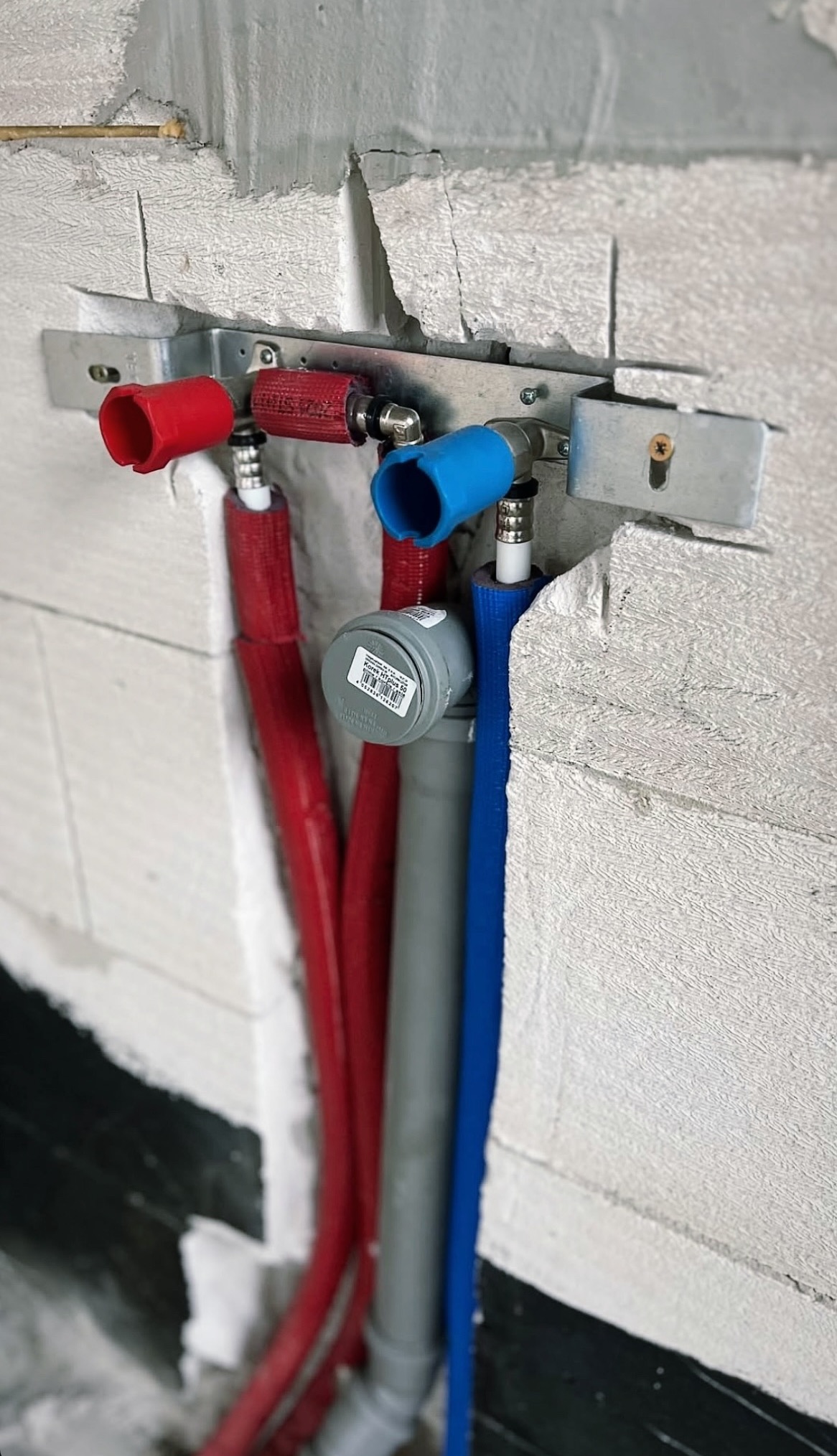 Instalacja wodno-kanalizacyjna w surowym stanie: rury PEX w izolacji (czerwona, niebieska) i szara rura kanalizacyjna, zamocowane do ściany z betonu komórkowego.