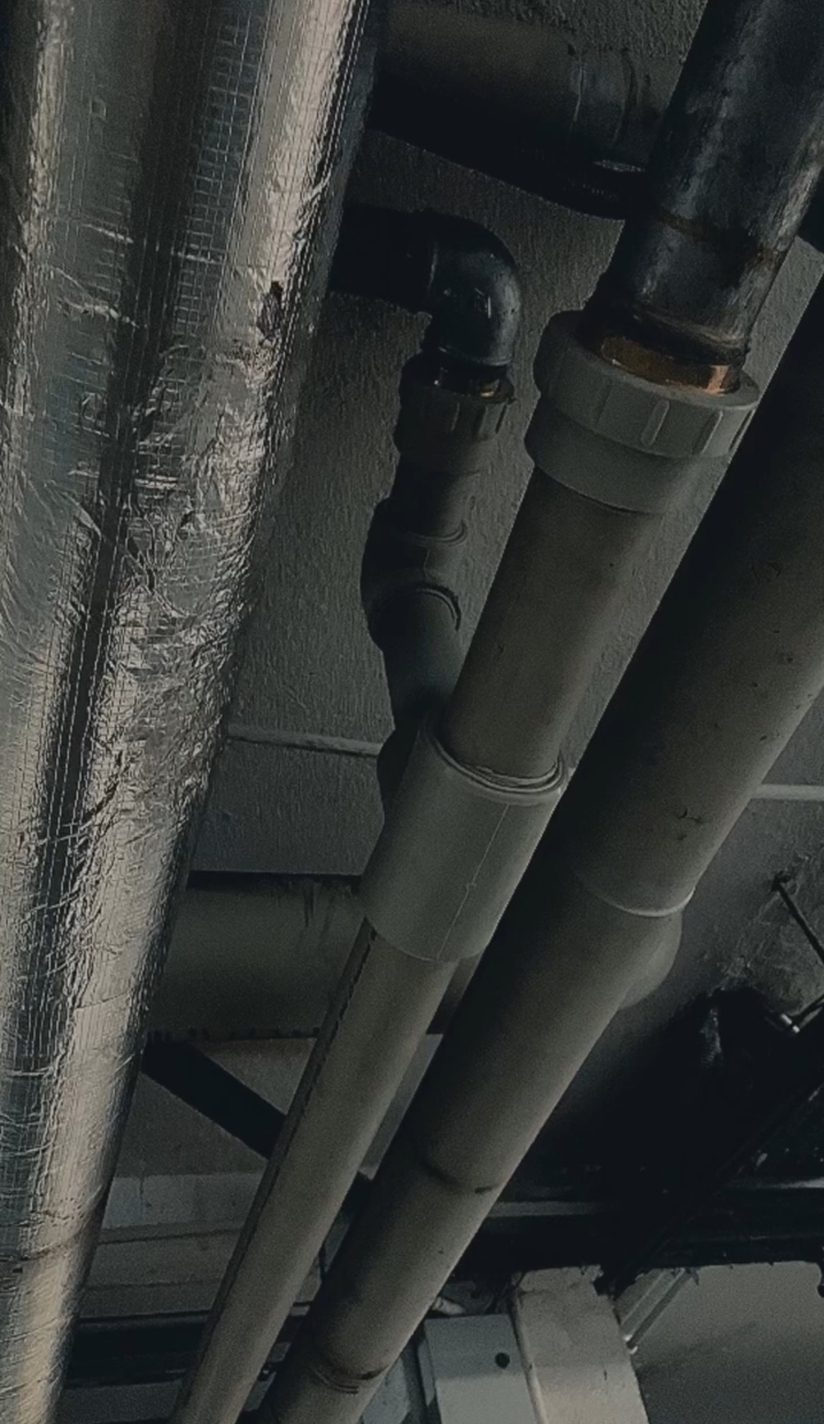 Szare rury kanalizacyjne i rura z izolacją termiczną, widoczne pod kątem od dołu, w pomieszczeniu o surowym wykończeniu. Elementy instalacji połączone złączkami.