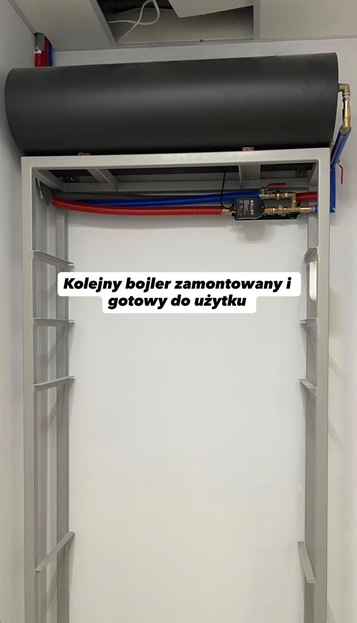Zamontowany, poziomy bojler w metalowej ramie, z widocznymi rurami doprowadzającymi wodę. Instalacja w pomieszczeniu o białych ścianach i suficie.