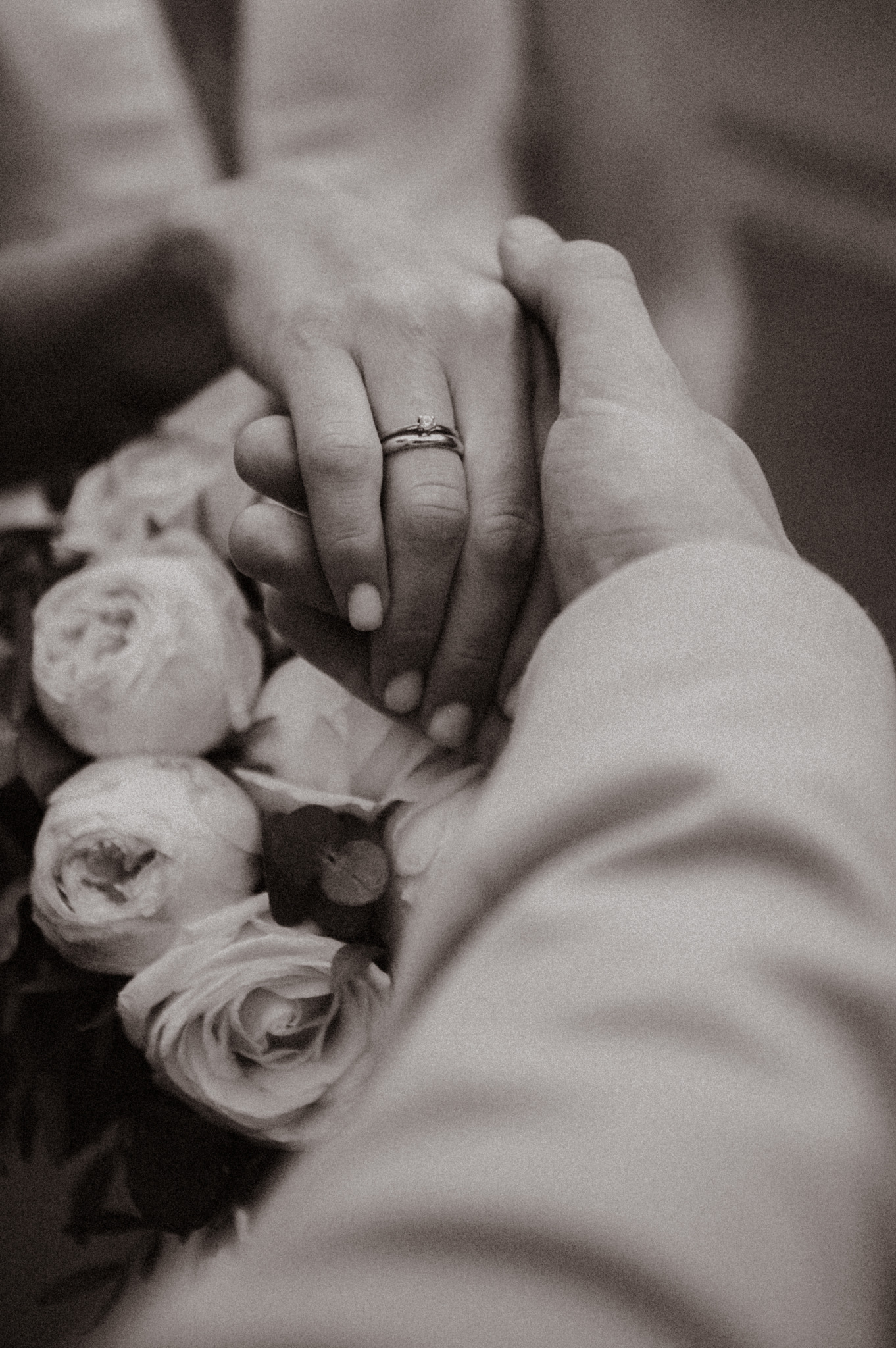 Zbliżenie dłoni pary młodej z pierścionkiem zaręczynowym na tle bukietu ślubnego w odcieniach bieli i kremu, w sepii. Delikatne światło podkreśla detale.