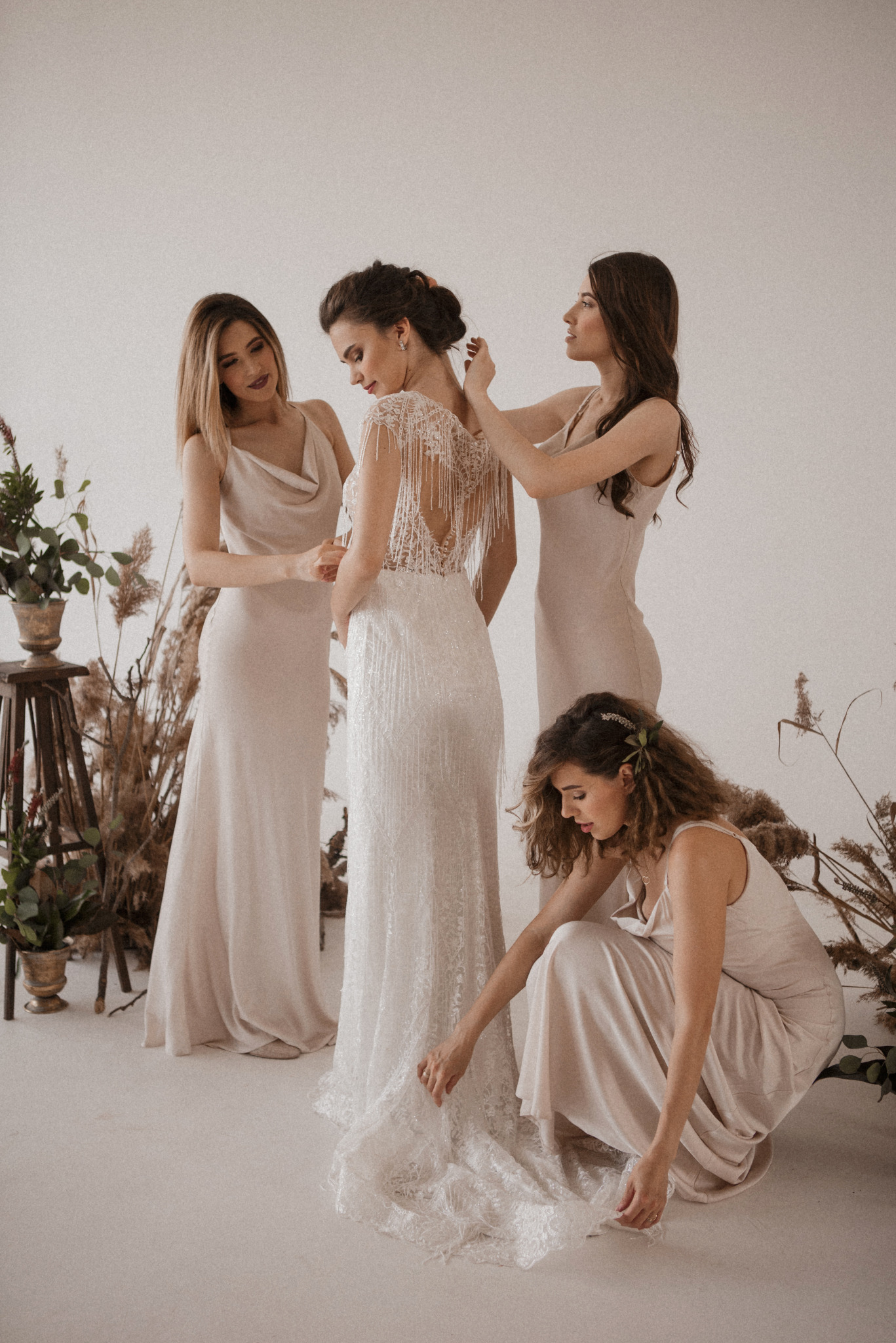 Panna młoda w sukni ślubnej z odkrytymi plecami, otoczona druhnami w beżowych sukienkach, poprawiającymi jej strój. Dekoracje z suszonych traw w tle.