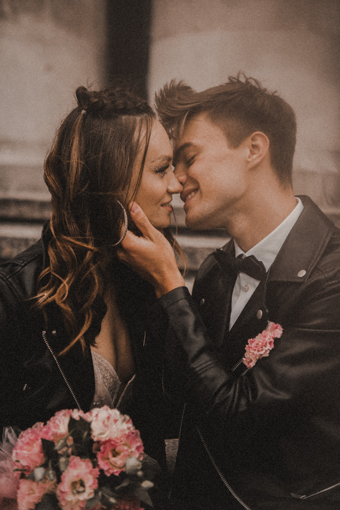 Para młoda w skórzanych kurtkach, zbliżona do pocałunku, panna młoda z brokatem na powiekach, bukiet różowych kwiatów, stylizacja rockowa, fotografia ślubna.