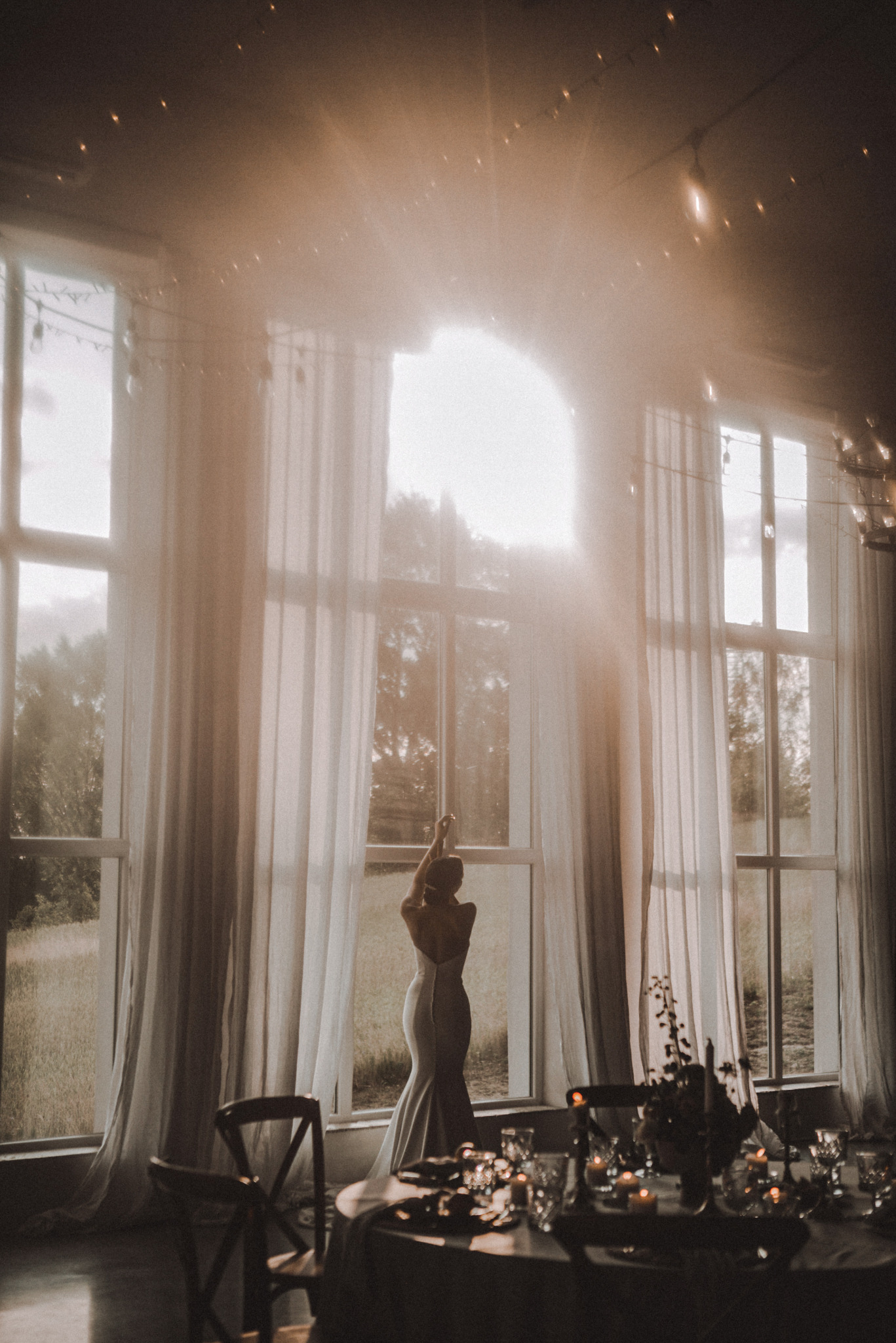 Panna młoda w sukni stoi tyłem przy oknie z firanami, światło słoneczne wpada do sali weselnej z dekoracjami stołu i krzesłami. Romantyczna atmosfera.