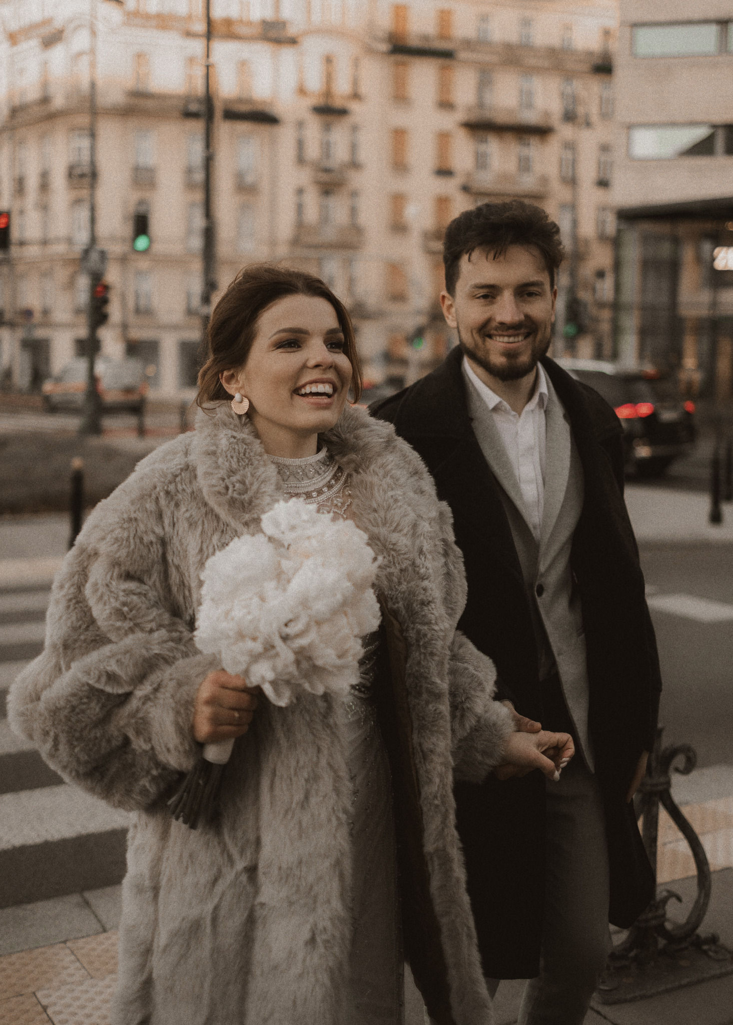 Uśmiechnięta para młoda w miejskiej scenerii. Panna młoda w futrze trzyma bukiet białych kwiatów, obok niej pan młody w eleganckim płaszczu. W tle budynki i sygnalizacja świetlna.