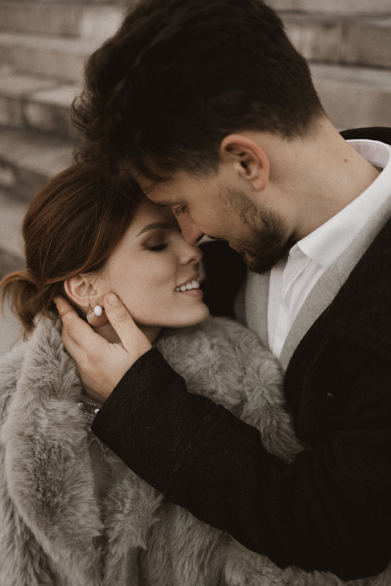 Delikatne zbliżenie na parę w objęciach, panna młoda w futrze, subtelny uśmiech, ciepłe światło, intymna chwila, eleganckie kolczyki, stonowana kolorystyka.