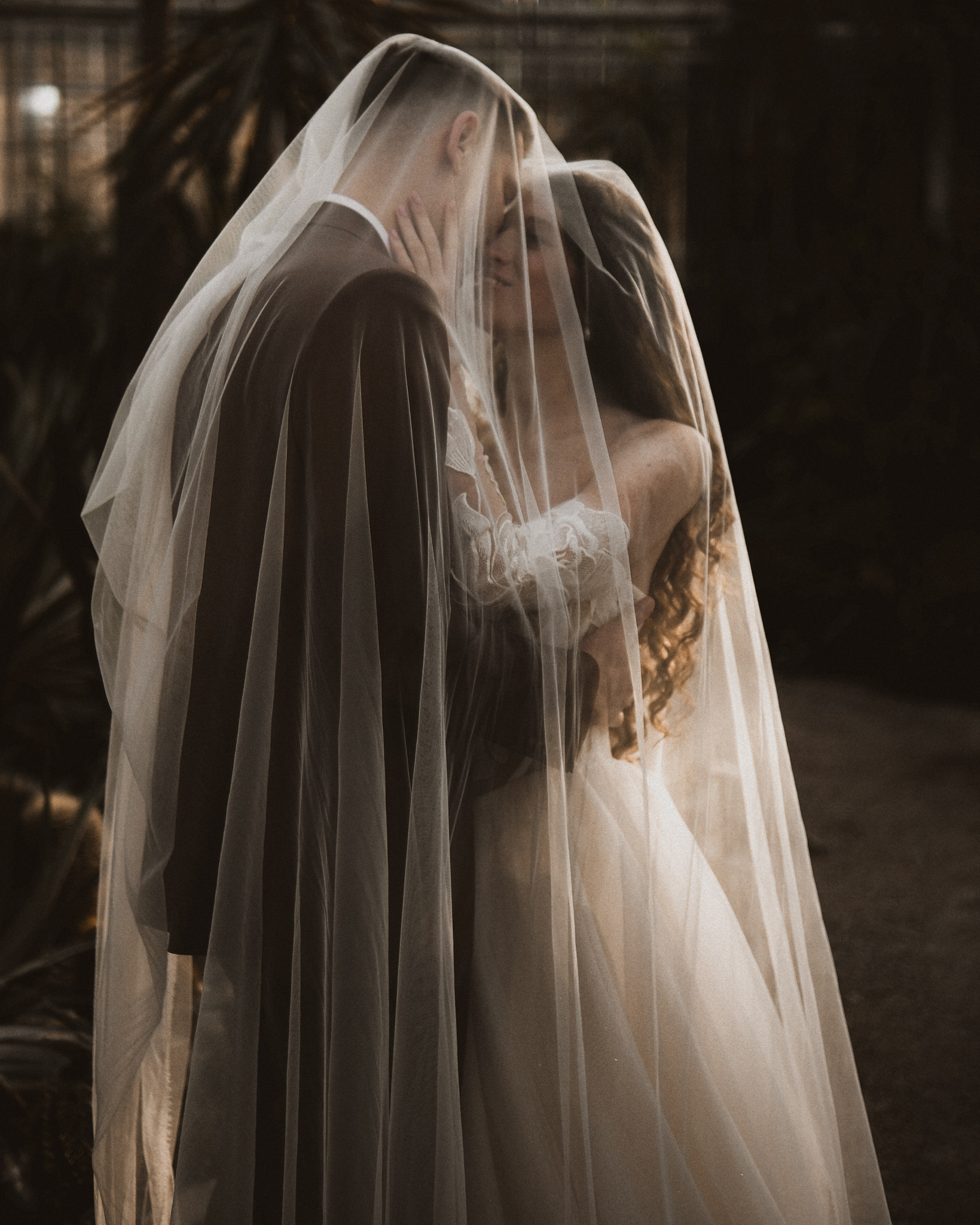 Para młoda pod welonem w intymnym uścisku. Delikatne światło podkreśla romantyczny nastrój. Widoczne fragmenty sukni ślubnej i garnituru. Tło rozmyte, naturalne.