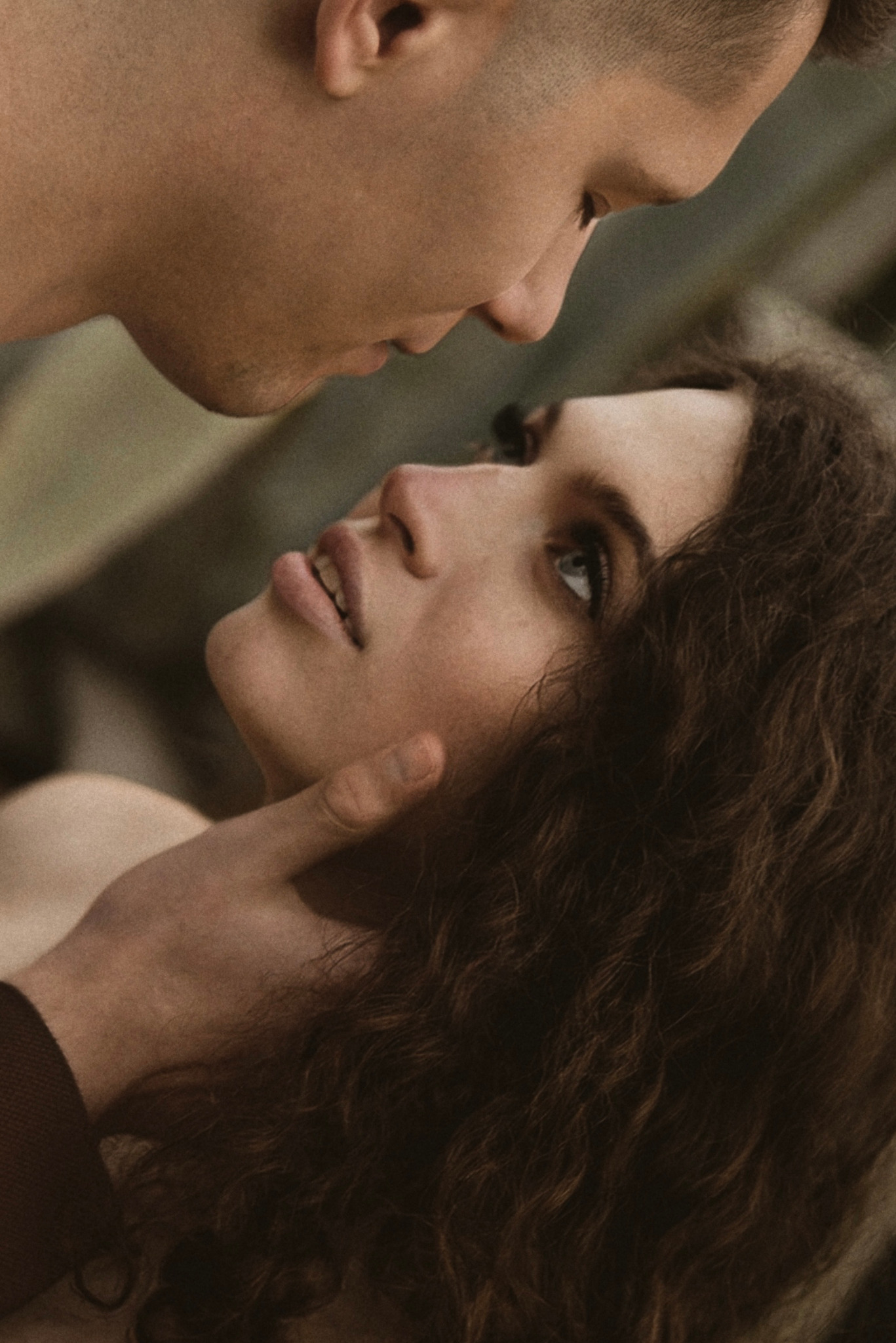 Delikatne zbliżenie na parę: on patrzy z góry, dotykając jej twarzy. Ona spogląda w górę, z widocznymi kręconymi włosami i subtelnym makijażem. Ciepłe, naturalne światło.