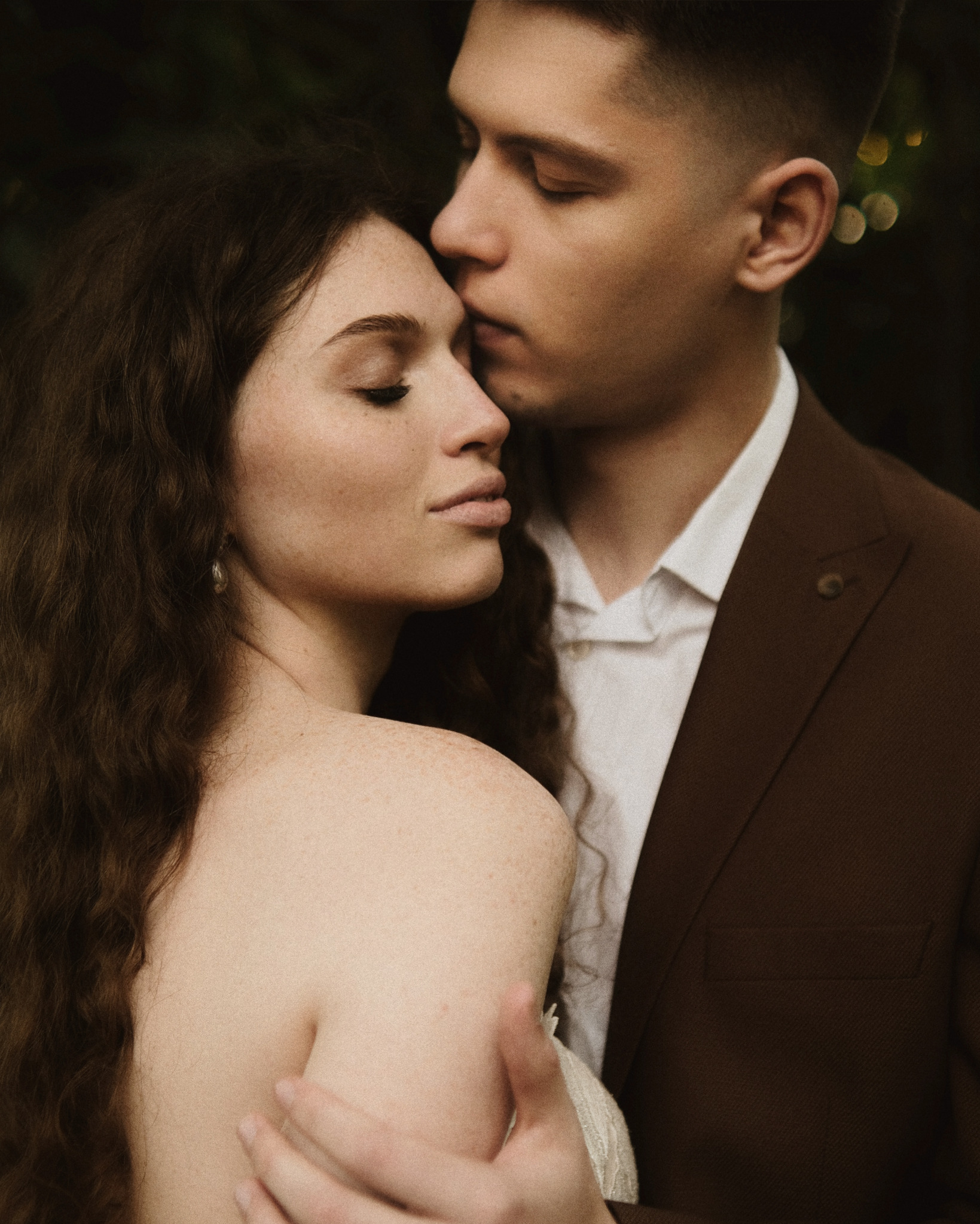 Delikatne ujęcie pary: mężczyzna w brązowym garniturze całuje kobietę w czoło. Romantyczna fotografia ślubna z subtelnym oświetleniem i naturalną kolorystyką.