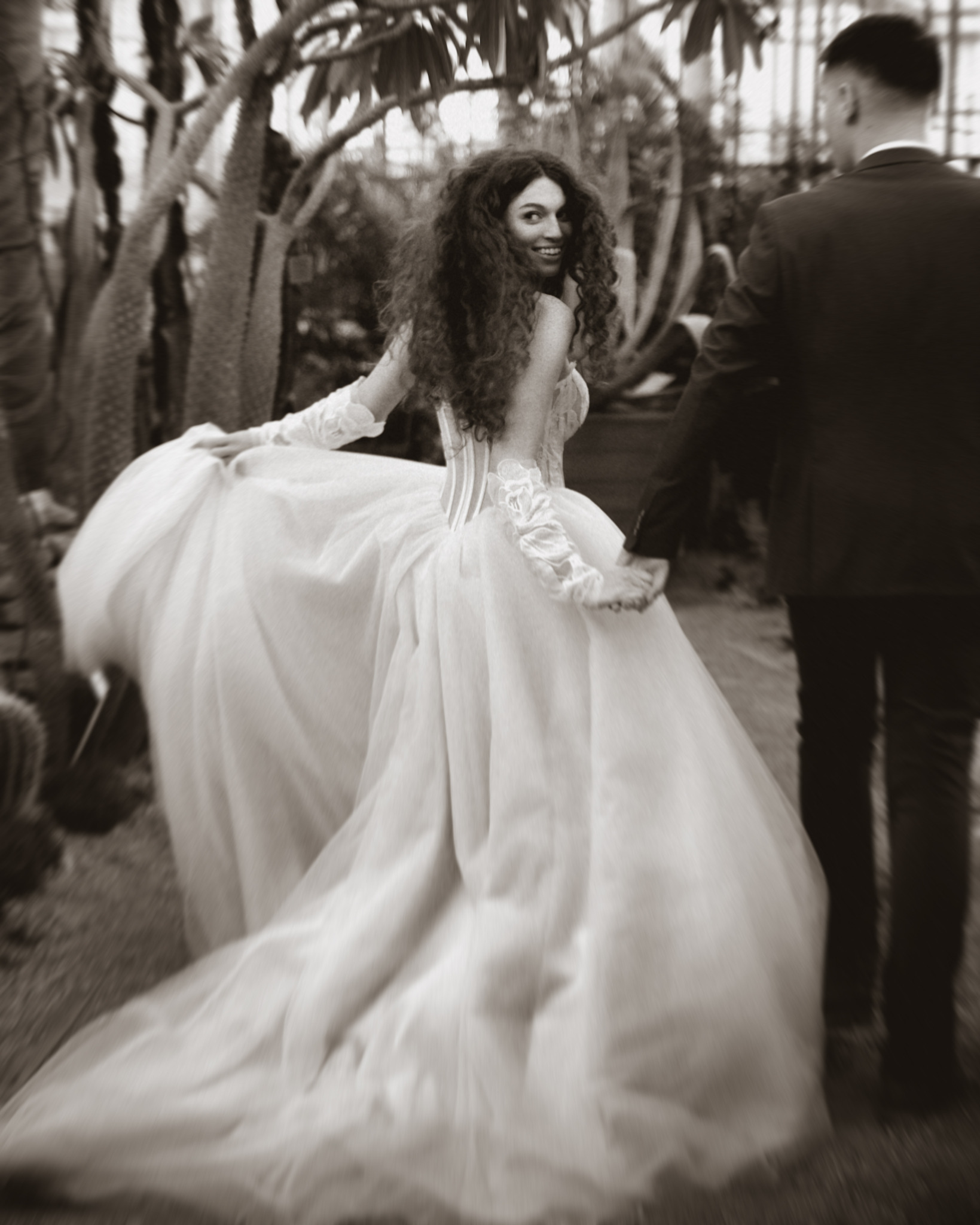 Sepia: Panna młoda w koronkowej sukni spogląda przez ramię, trzymając dłoń pana młodego na tle egzotycznej roślinności. Romantyczna sesja w botanicznym ogrodzie.