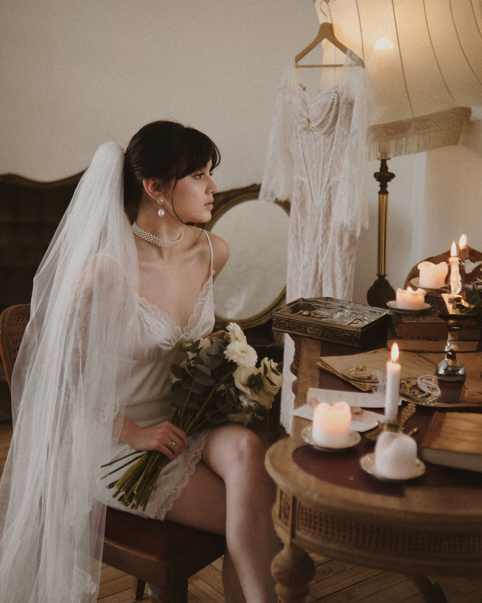 Panna młoda w bieliźnie i welonie siedzi przy stoliku ze świecami, trzyma bukiet. W tle suknia ślubna i lustro. Klimatyczne, intymne ujęcie przedślubne.
