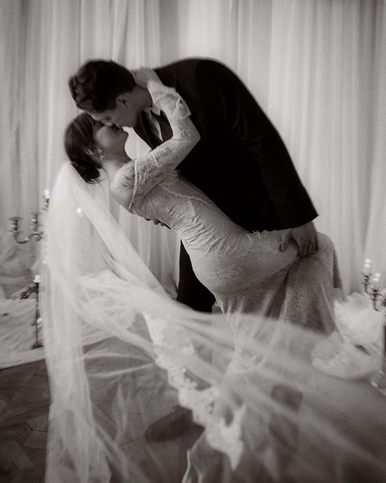 Czarno-białe zdjęcie ślubne: Panna Młoda w koronkowej sukni i welonie, pochylona w pocałunku z Panem Młodym w ciemnym garniturze, świeczniki w tle.