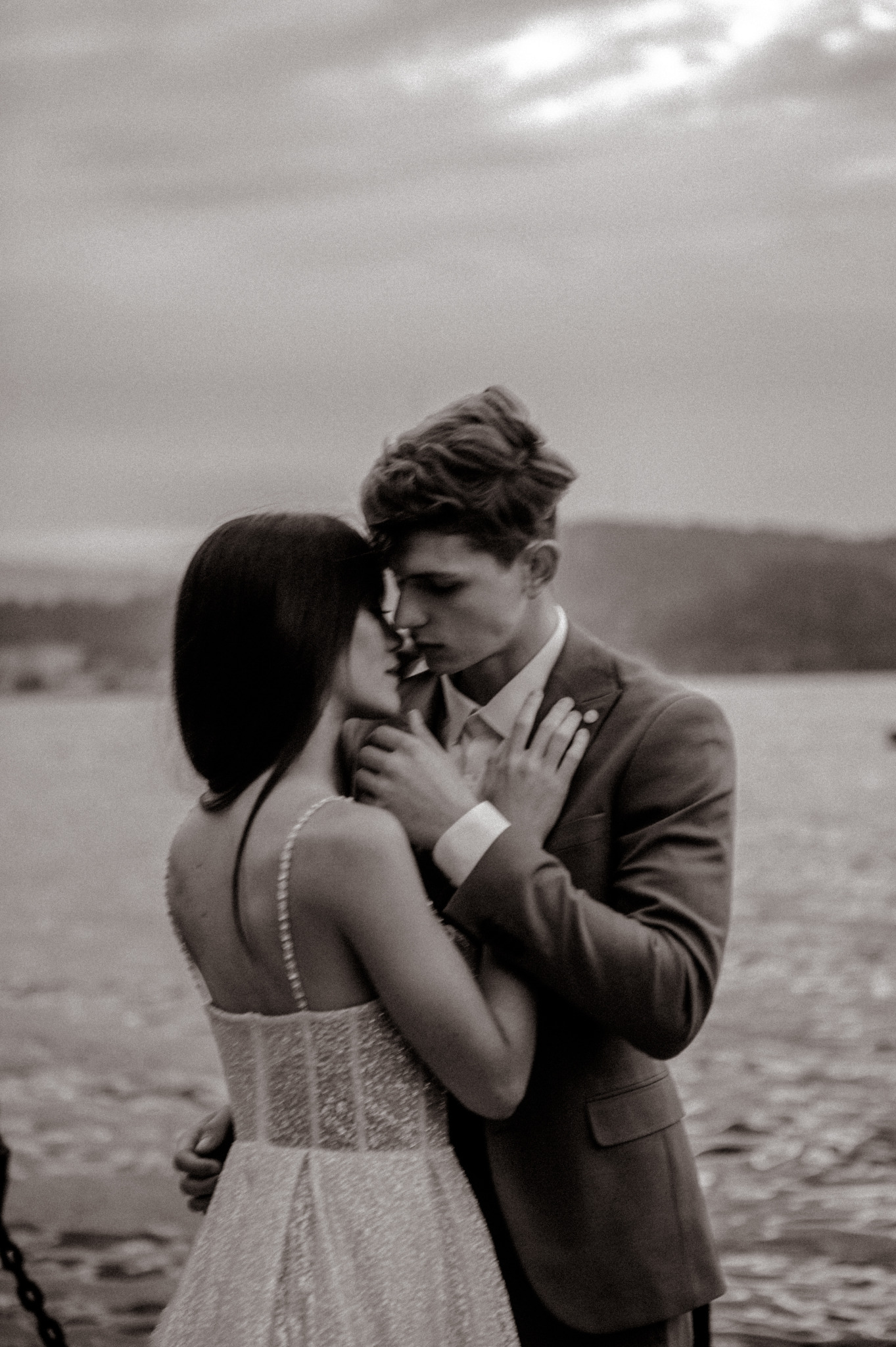 Sepiowe zbliżenie: para młoda w objęciach na tle jeziora. Ona w sukni z delikatnymi ramiączkami, on w garniturze. Romantyczny moment, naturalne światło.