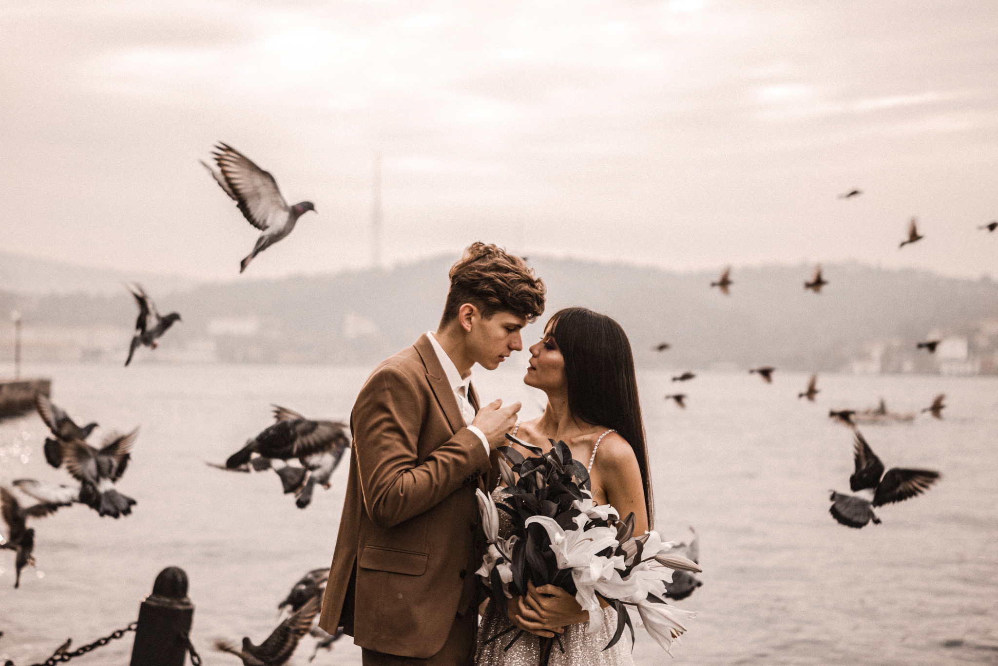Zakochana para młoda w objęciach, z bukietem lilii na tle morza i stada gołębi w locie. Ujęcie w ciepłej tonacji, z delikatnym rozmyciem tła.