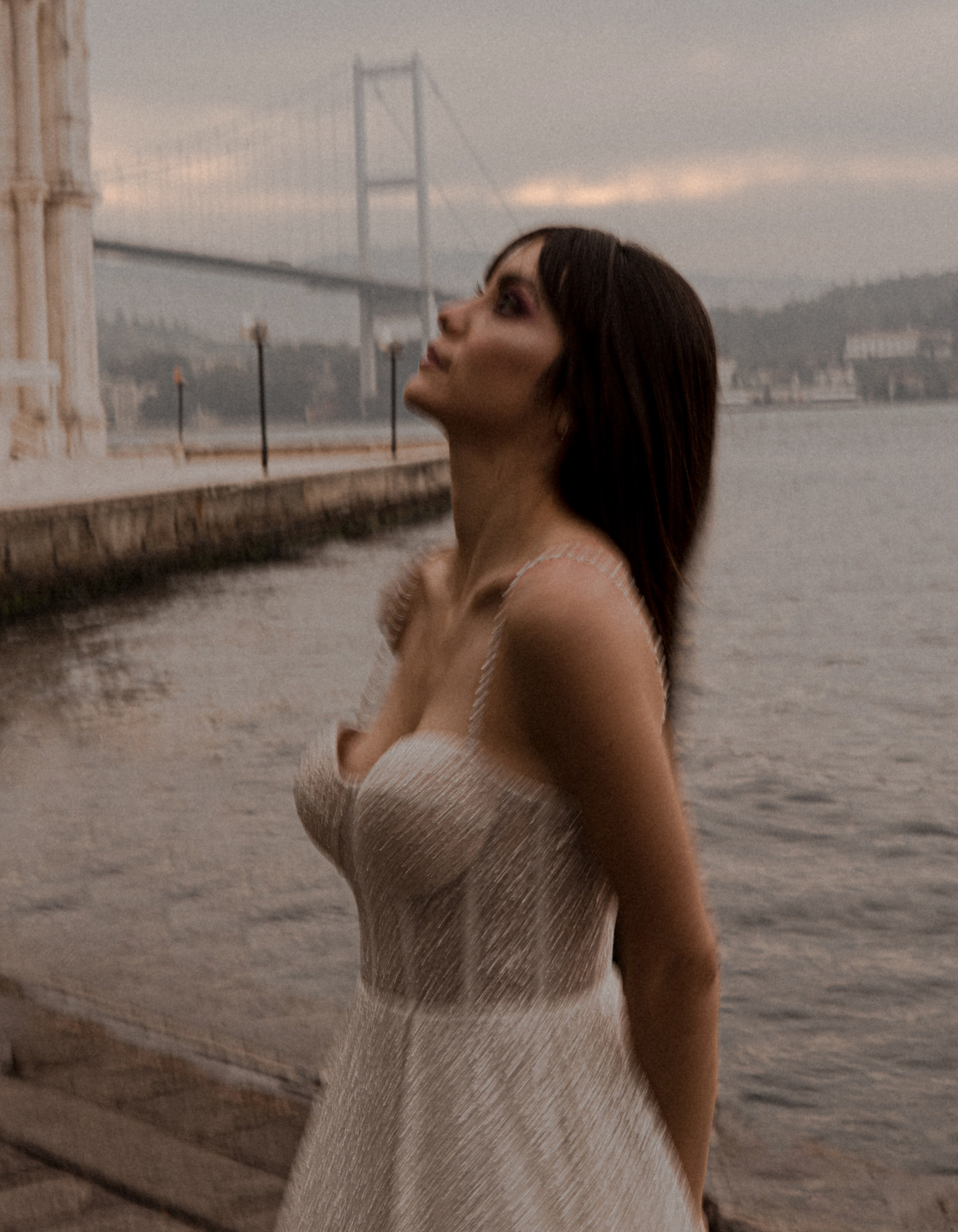 Panna młoda w sukni ślubnej spogląda w górę na tle mostu w Warszawie. Delikatne, rozproszone światło podkreśla subtelność sukni i makijażu.