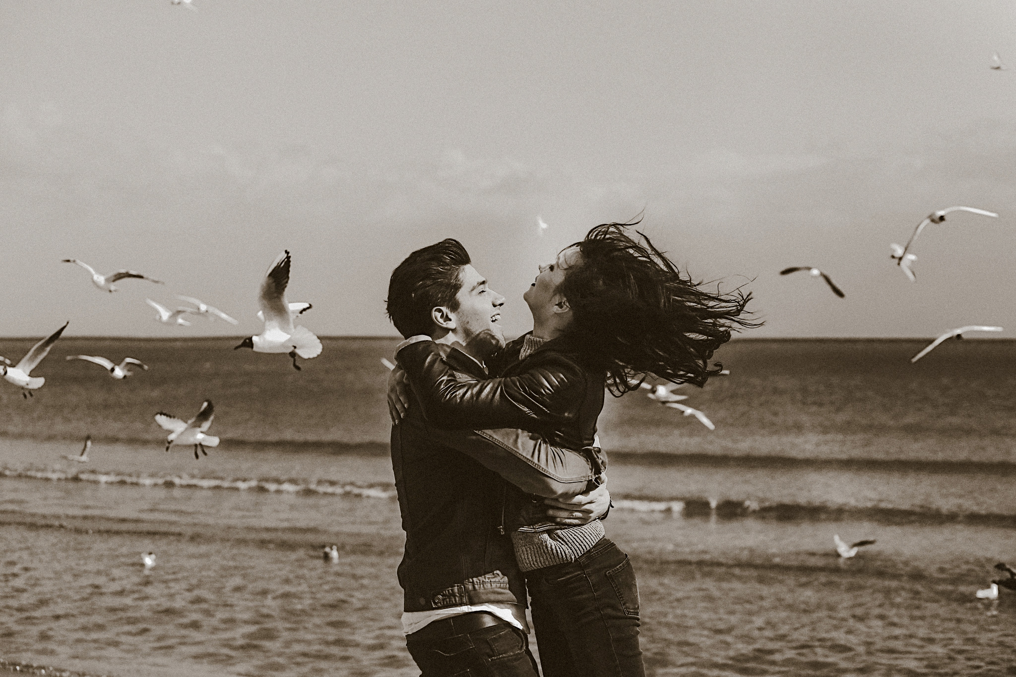 Sepia fotografia ślubna: para w objęciach na plaży, włosy rozwiane wiatrem, w tle morze i stado mew w locie. Romantyczna sesja plenerowa.