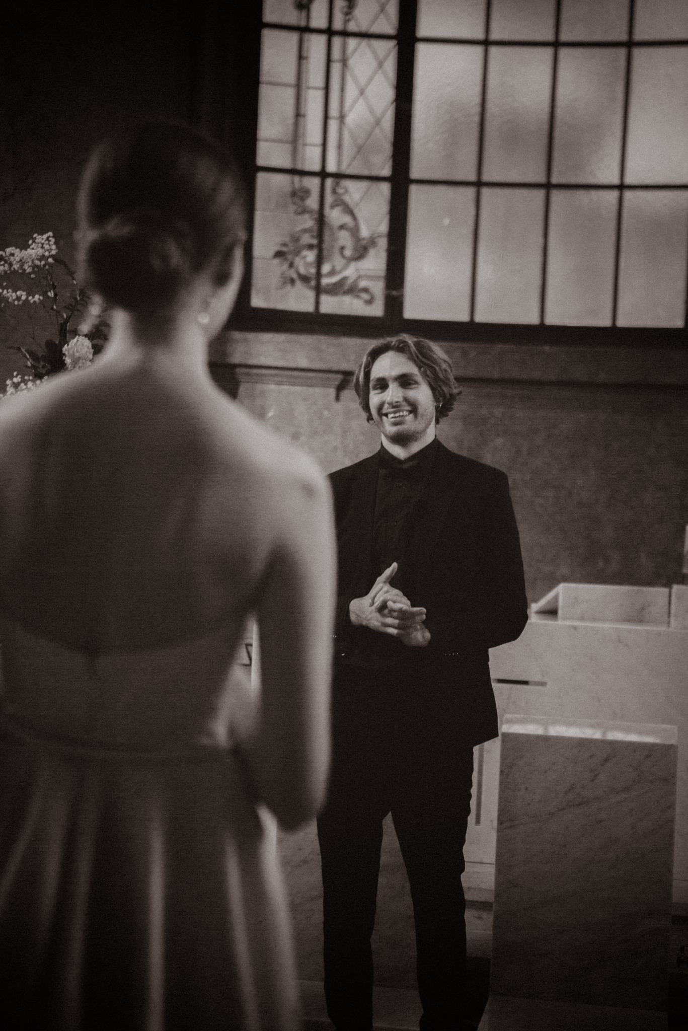 Czarno-białe zdjęcie z uroczystości ślubnej: uśmiechnięty pan młody w ciemnym garniturze, widziany zza pleców panny młodej w jasnej sukni, w tle okno.