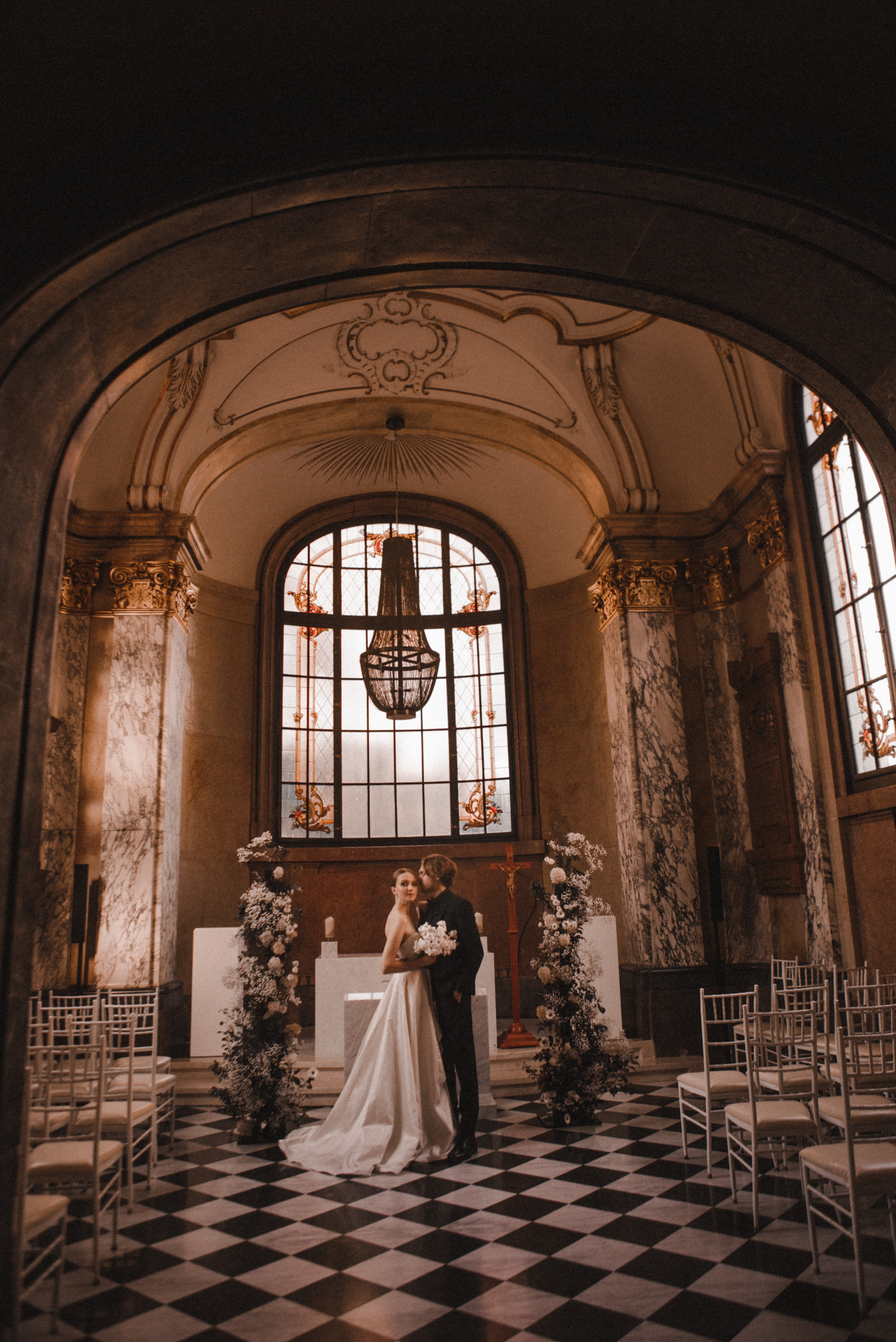 Elegancka sesja ślubna w zabytkowym wnętrzu: para młoda w objęciach na tle marmurowych kolumn, okien witrażowych i szachownicy na podłodze. Romantyczna atmosfera.
