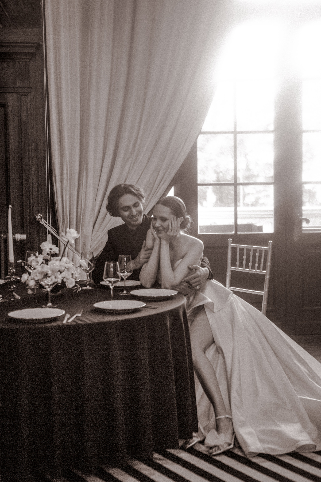 Sepiowe zdjęcie: uśmiechnięta para młoda siedzi przy stole z kwiatami i zastawą, w tle okno z jasnym światłem. Romantyczna atmosfera, eleganckie wnętrze.