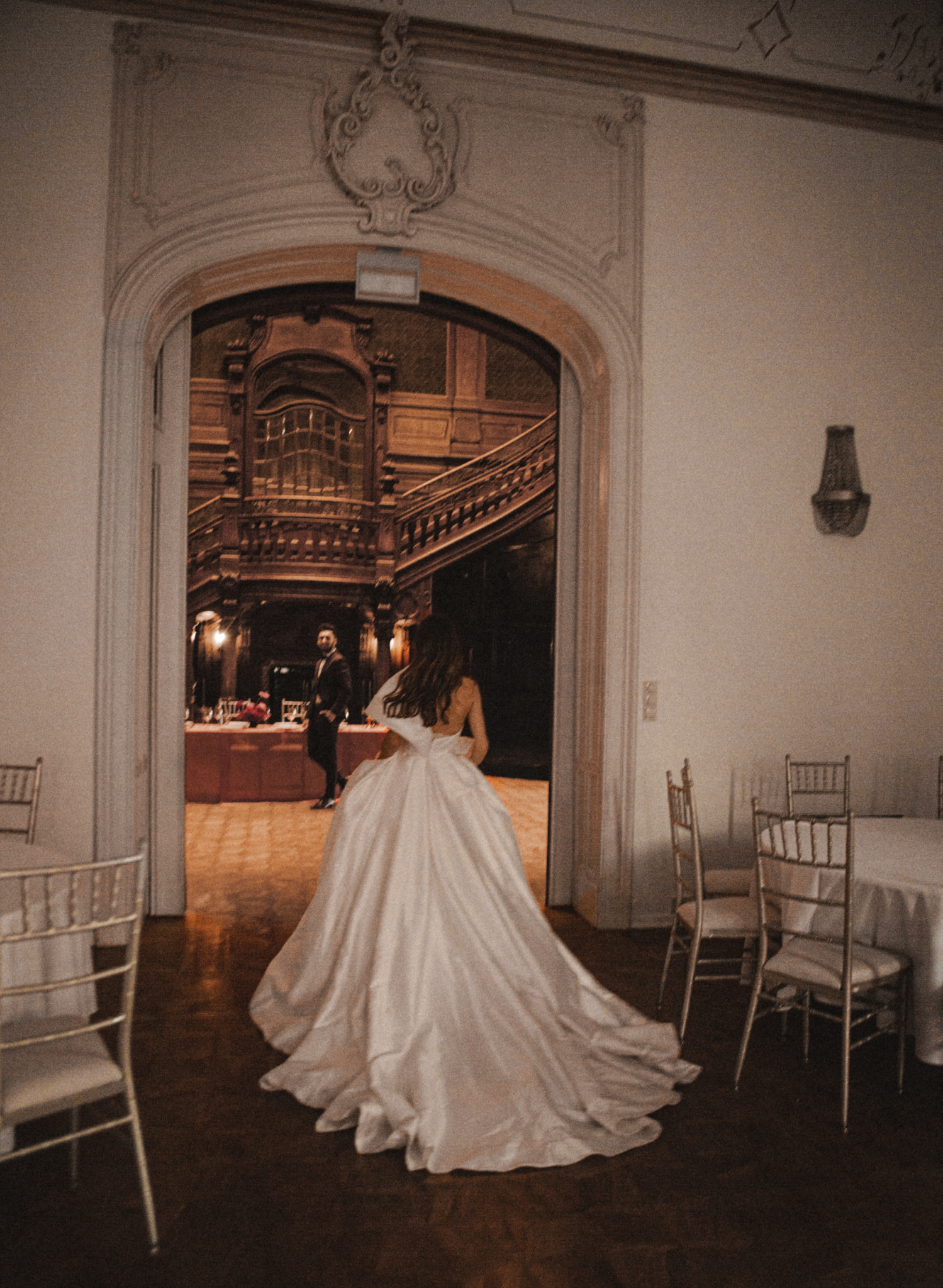 Panna młoda w sukni przechodzi przez ozdobny portal do sali balowej z drewnianą klatką schodową i stołem z kwiatami, gdzie czeka pan młody. Eleganckie krzesła i stoły w tle.