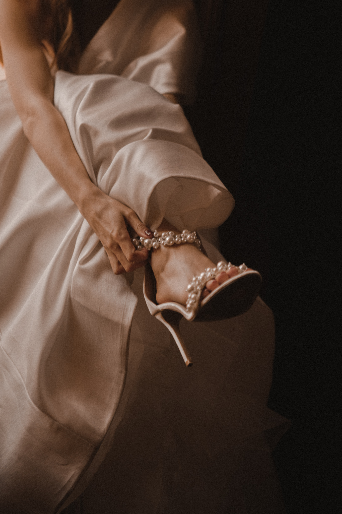 Panna młoda w satynowej sukni zapina elegancki, perłowy sandał na obcasie. Delikatne światło podkreśla detale sukni i buta na ciemnym tle.