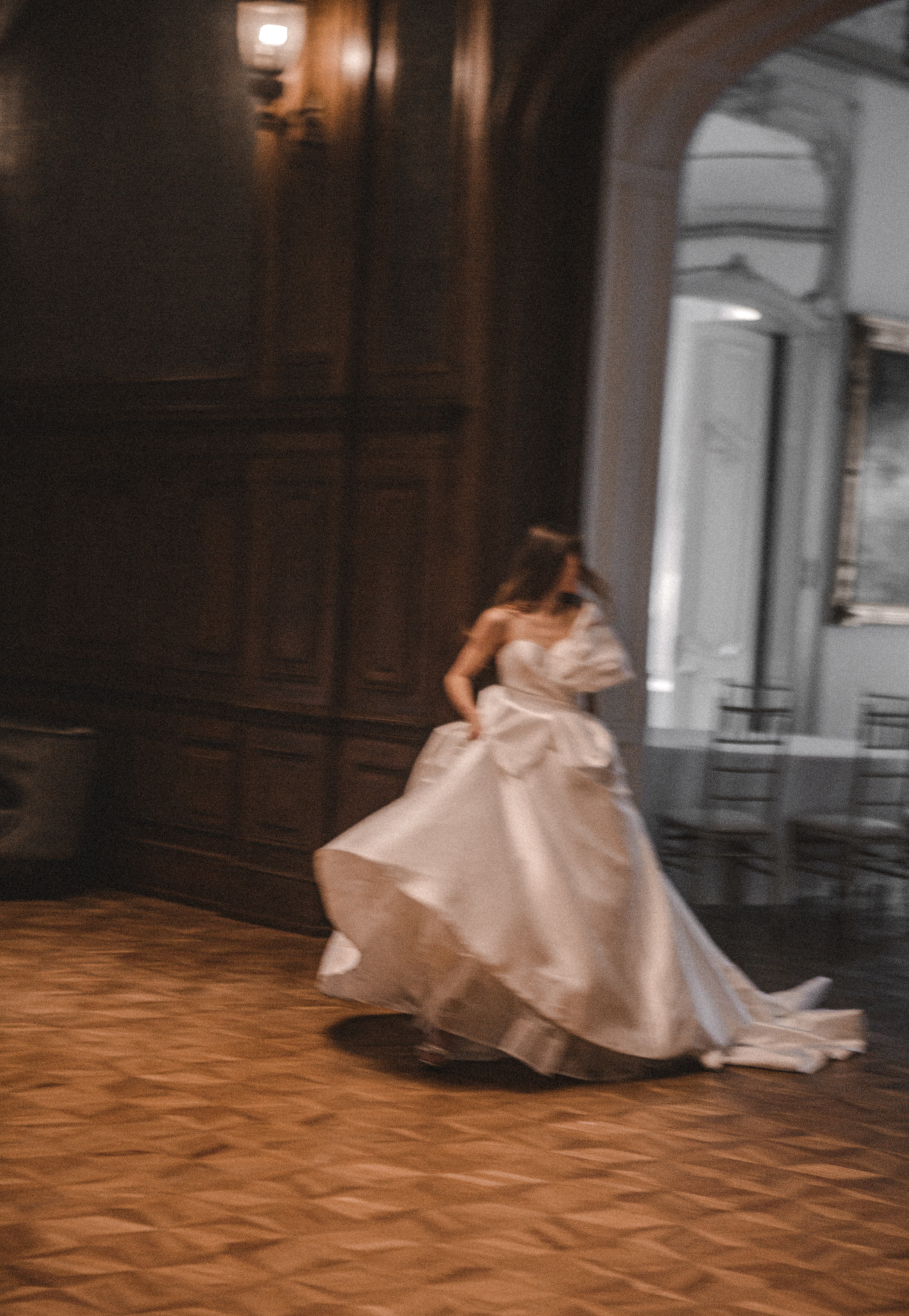 Panna młoda w białej sukni biegnie po parkiecie w zabytkowym wnętrzu z drewnianą boazerią i łukowym przejściem. Rozmycie ruchu dodaje dynamiki.