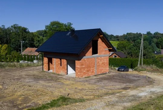 Niedokończony, parterowy dom z czerwonej cegły z ciemnym dachem na tle zieleni i błękitnego nieba. Widoczny samochód osobowy w oddali. Stan surowy otwarty.