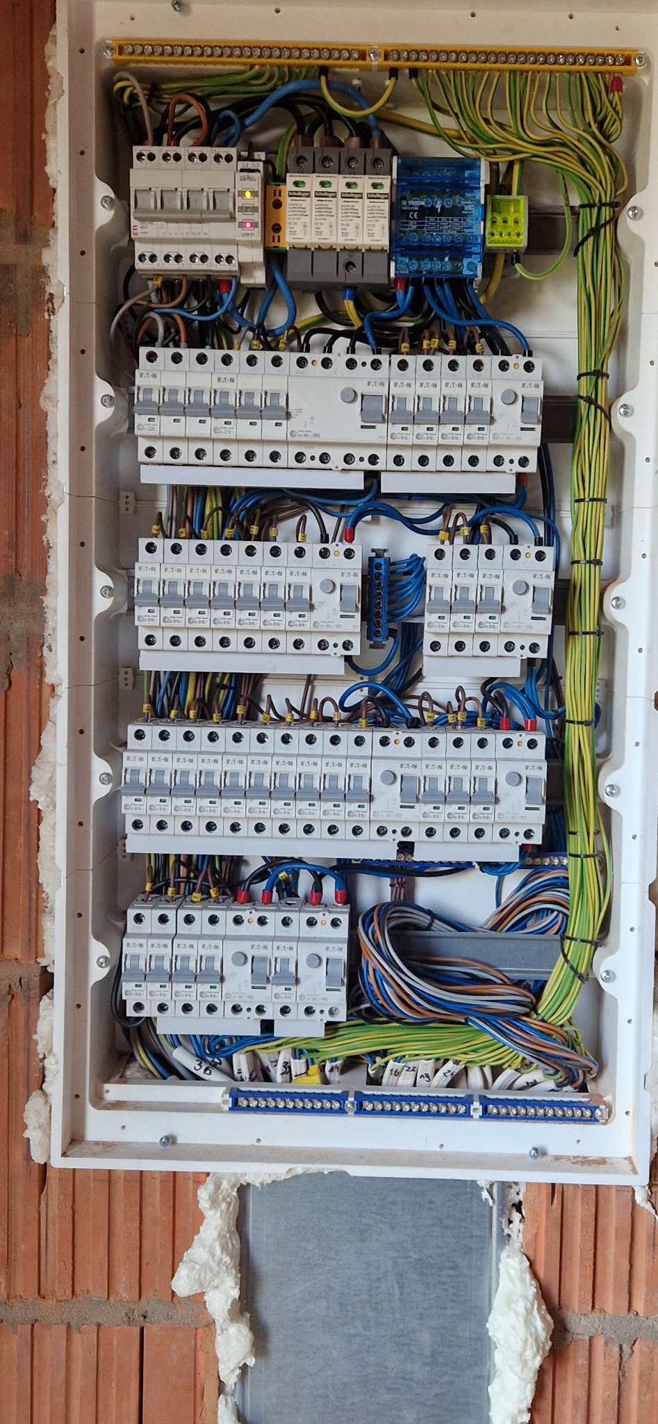Rozdzielnica elektryczna w białej obudowie, zamontowana na ceglanej ścianie, z widocznymi zabezpieczeniami, przewodami i elementami instalacji. Widoczne pianowanie wokół obudowy.