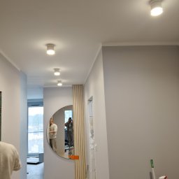 Remonty Wykończenia pod klucz od A do Z - Jasne wnętrze po remoncie: gładkie, szare ściany, okrągłe lustro z drewnianą dekoracją, sufit z oświetleniem punktowym. Widoczne narzędzia malarskie i odzież robocza.