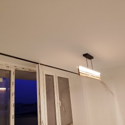 Remonty Wykończenia pod klucz od A do Z - Nowoczesna lampa wisząca z drewnianym akcentem oświetla białe wnętrze z oknem. Widoczne ślady po instalacji elektrycznej na ścianie.